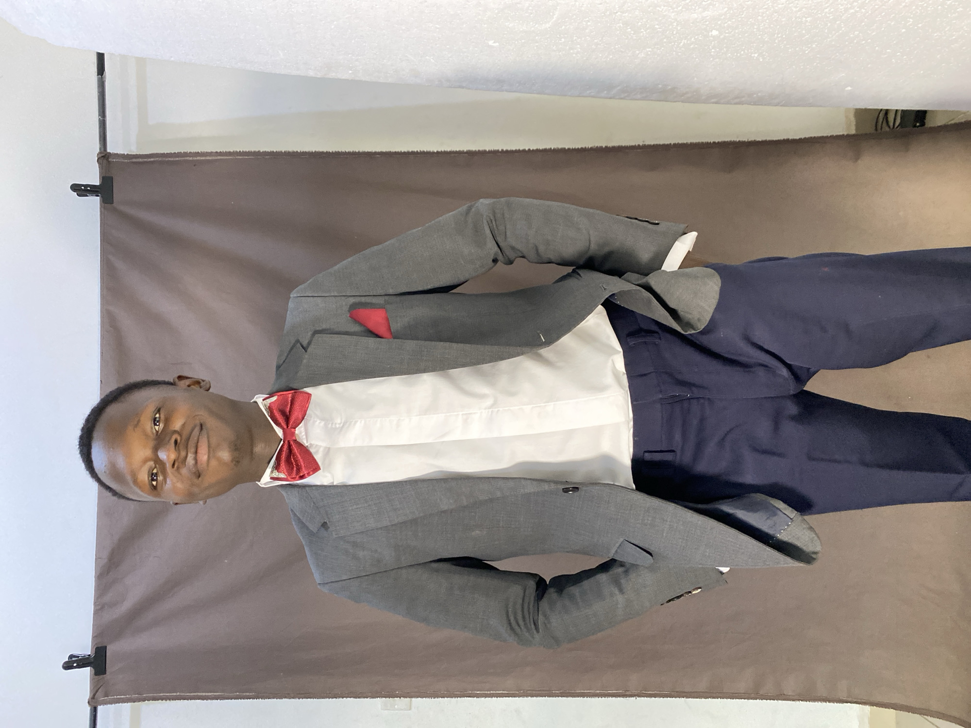 A young Black male stands in a studio, wearing a gray suit jacket, white shirt, navy trousers, a red bowtie, and a red pocket square. The background consists of a brown fabric backdrop and a white textured surface on the right. The lighting is even, and the subject is looking at the camera with a slight smile. The image is framed from the chest up.