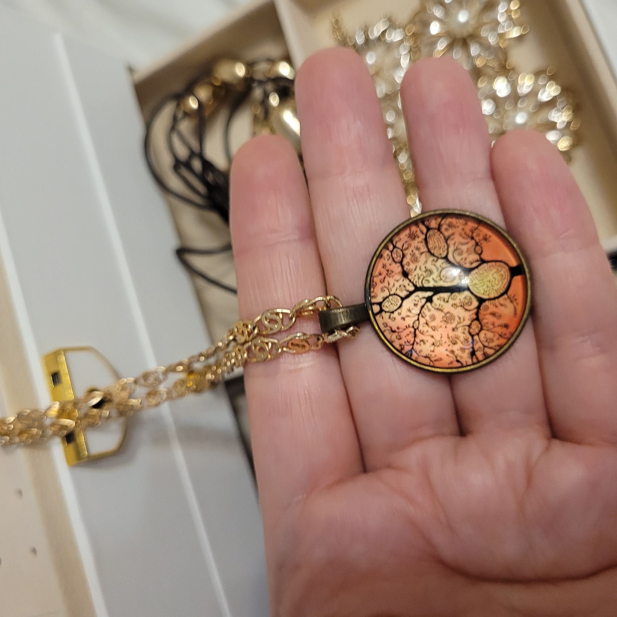 A person's hand holds up a gold-toned necklace featuring a distinctive round pendant. The pendant showcases an intricate, stylized design resembling a branching tree or a neural network, rendered in black on a vibrant orange and gold background, and framed by a bronze-colored bezel. The necklace chain is also gold-colored, with an intertwined link pattern.
The hand, with fair skin, gently presents the pendant. In the blurred background, a light-colored jewelry storage box or tray is visible, containing other out-of-focus gold-toned jewelry items that produce soft bokeh effects. The setting appears to be indoors, likely a personal space where jewelry is stored, and the activity suggests the person is examining or displaying the necklace. No visible text is present.