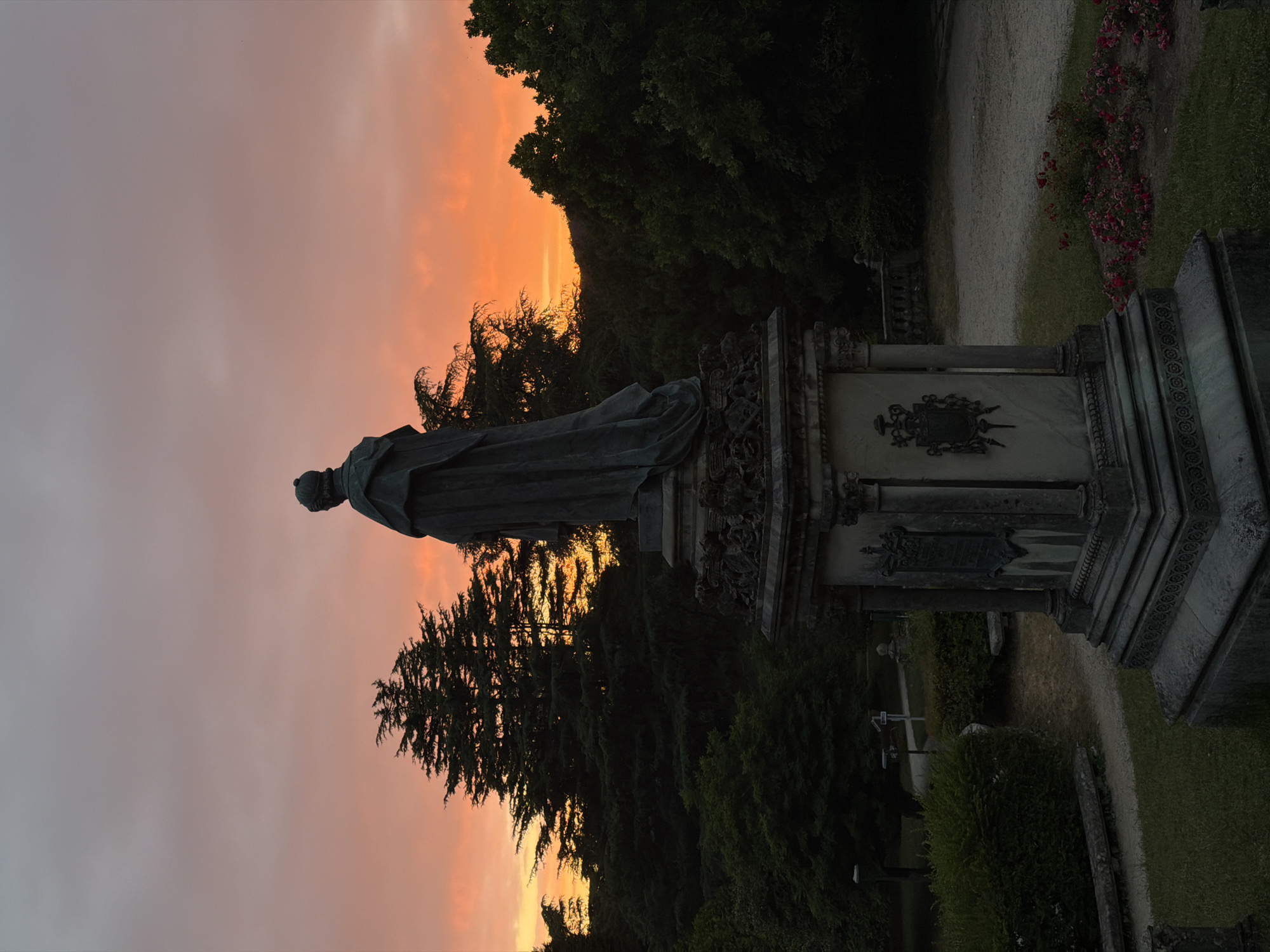 The image depicts a monumental statue in a cemetery, silhouetted against a vibrant sunset. The statue, made of dark material, stands on a tiered pedestal adorned with decorative carvings. The figure is draped in what appears to be a robe, with its back to the viewer, facing the fiery orange and pink sky. Evergreen trees and lush foliage form a dark backdrop, with hills visible in the distance. The foreground shows manicured grass, pathways, and a patch of red flowers. The scene suggests a peaceful and somber atmosphere, characteristic of a cemetery at dusk. The location is not Santiago De Compostela, Spain, as there are no visible landmarks or architectural elements that identify it as such.