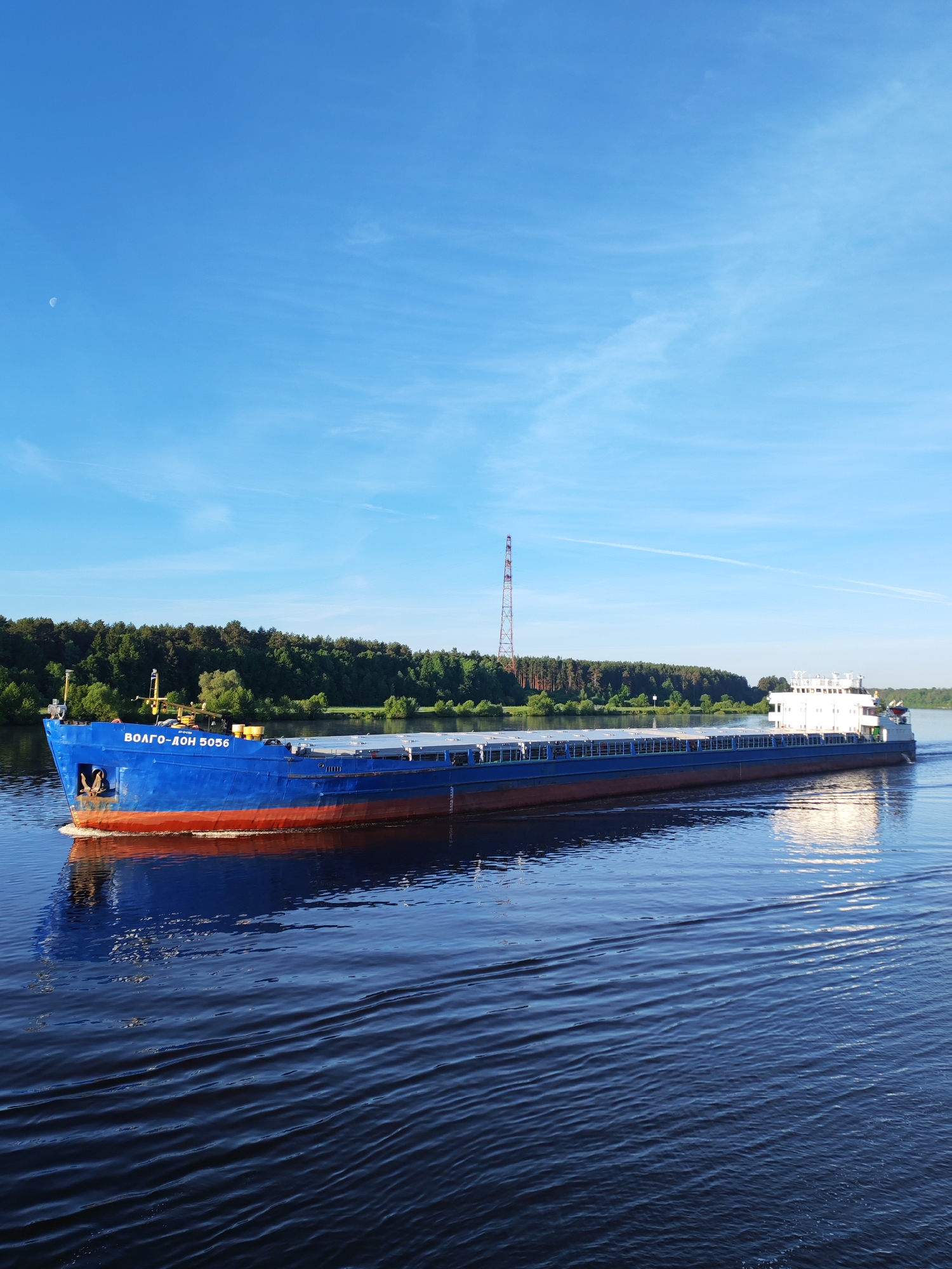 A large, blue-hulled cargo ship, bearing the name "ВОЛГО-ДОН 5056" in white Cyrillic script on its bow, traverses a wide river. The ship features a red anti-fouling paint line below the waterline and a white superstructure towards its stern. It generates a small bow wave and ripples across the dark blue water, which reflects the clear sky. The far bank is lined with dense green deciduous and coniferous trees under a bright blue sky with scattered wispy clouds. A distinct white crescent moon is visible in the upper left sky. On the distant tree-lined shore, a tall, red lattice transmission tower is prominent. The precise location in the city of Unknown, Unknown is undetermined, but the ship's name indicates operation on a waterway within the Volga-Don region. No individuals are visibly identifiable on the vessel or in the surrounding environment.