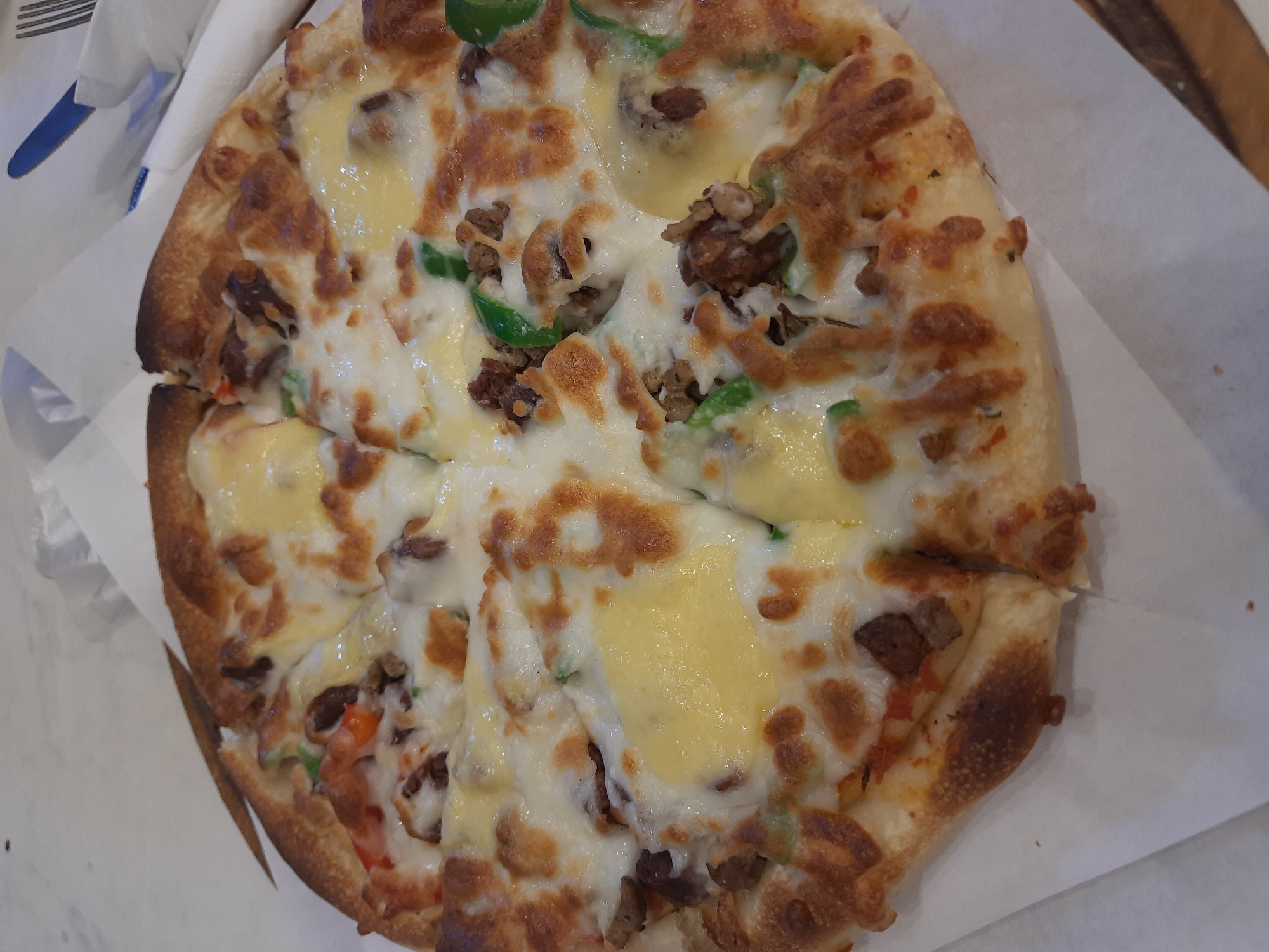 The image shows a partially eaten pizza cut into slices, placed on a white paper, possibly a napkin. The pizza has a thick crust, browned in some areas. The toppings include melted cheese (likely mozzarella and cheddar), small pieces of cooked meat (possibly beef), and green peppers. The pizza is presented against a neutral-colored background, suggesting it is on a table. Part of a blue and white object, possibly a napkin or dish, is visible in the upper-left corner.