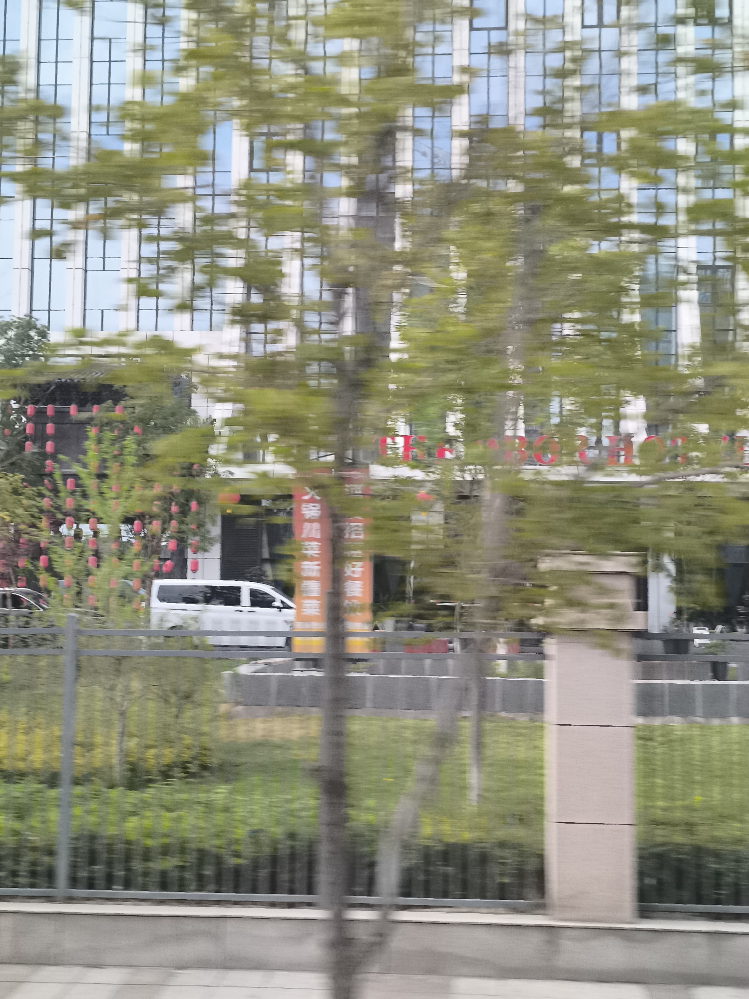 This is a photograph of a modern building with a glass facade in Ürümqi, China. The building's windows reflect the sky and surrounding trees. In the foreground, trees with green leaves are blurred due to motion, suggesting the photo was taken from a moving vehicle. A white van is parked near the building, and a fence runs along the side of the road. Red lanterns are strung on a tree branch, adding a decorative element. The overall scene depicts an urban environment with a mix of nature and architecture.