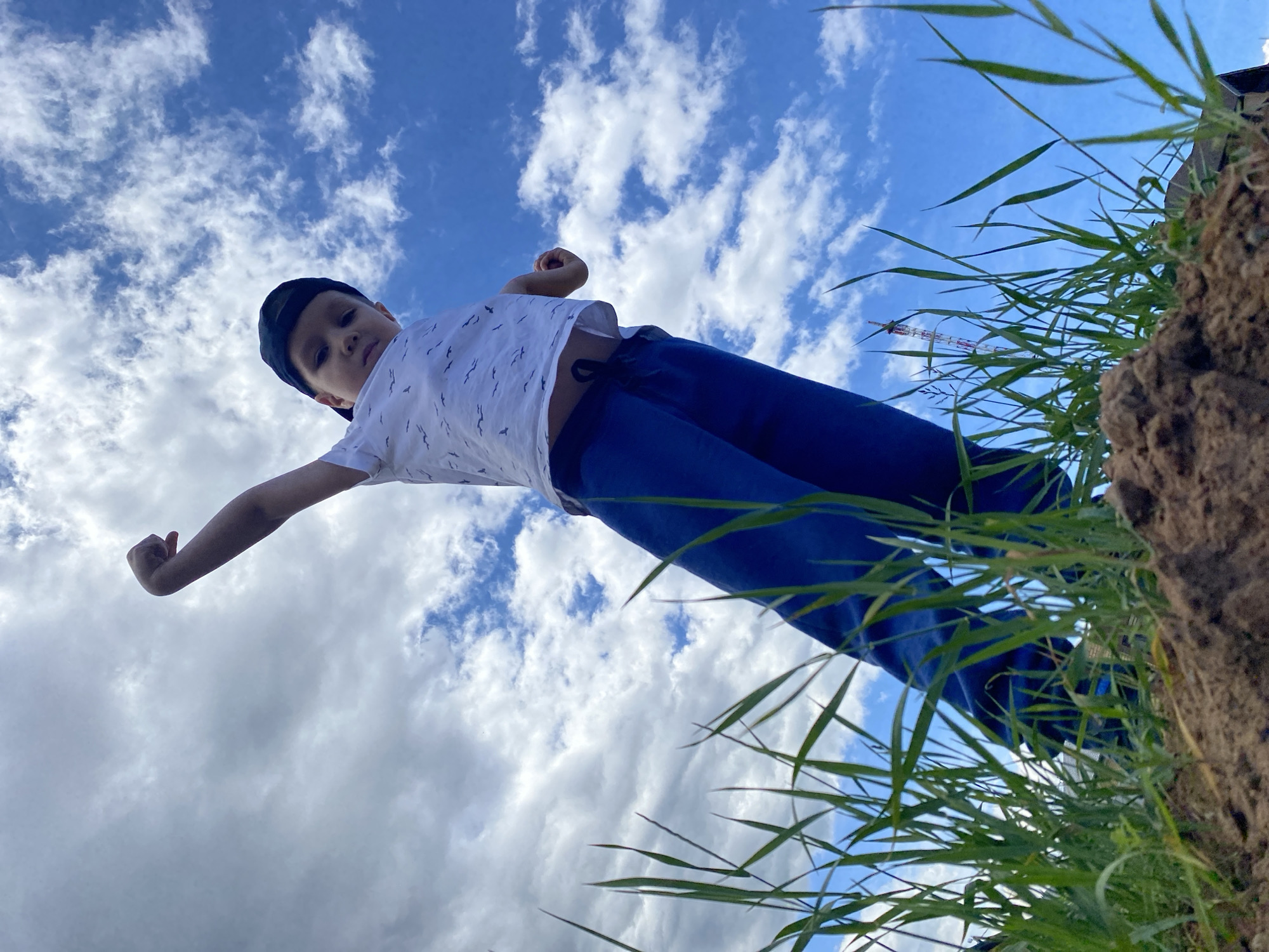 The image, taken from a low-angle perspective, depicts a young boy, approximately 5-8 years old, standing on elevated ground against a backdrop of a bright blue sky partially covered with white cumulus clouds. The boy is centrally positioned, looking downwards towards the camera with a neutral expression. He wears a white short-sleeved t-shirt patterned with small dark birds, dark blue athletic pants, and a dark-colored baseball cap worn towards the back of his head. His left arm is raised and bent at the elbow, with his hand lightly clenched, while his right arm is also bent with a partially visible clenched hand. The immediate foreground consists of a small mound of brown soil from which tall, green grass blades emerge, partially obscuring the lower view. In the distant background, visible above the grass and against the sky, a tall, slender lattice tower, featuring white and red stripes, is discernible. Hints of other structures, possibly building roofs, are also faintly visible through the grass. The scene takes place outdoors under daylight conditions, within the city of Chaikovsky, Russia.