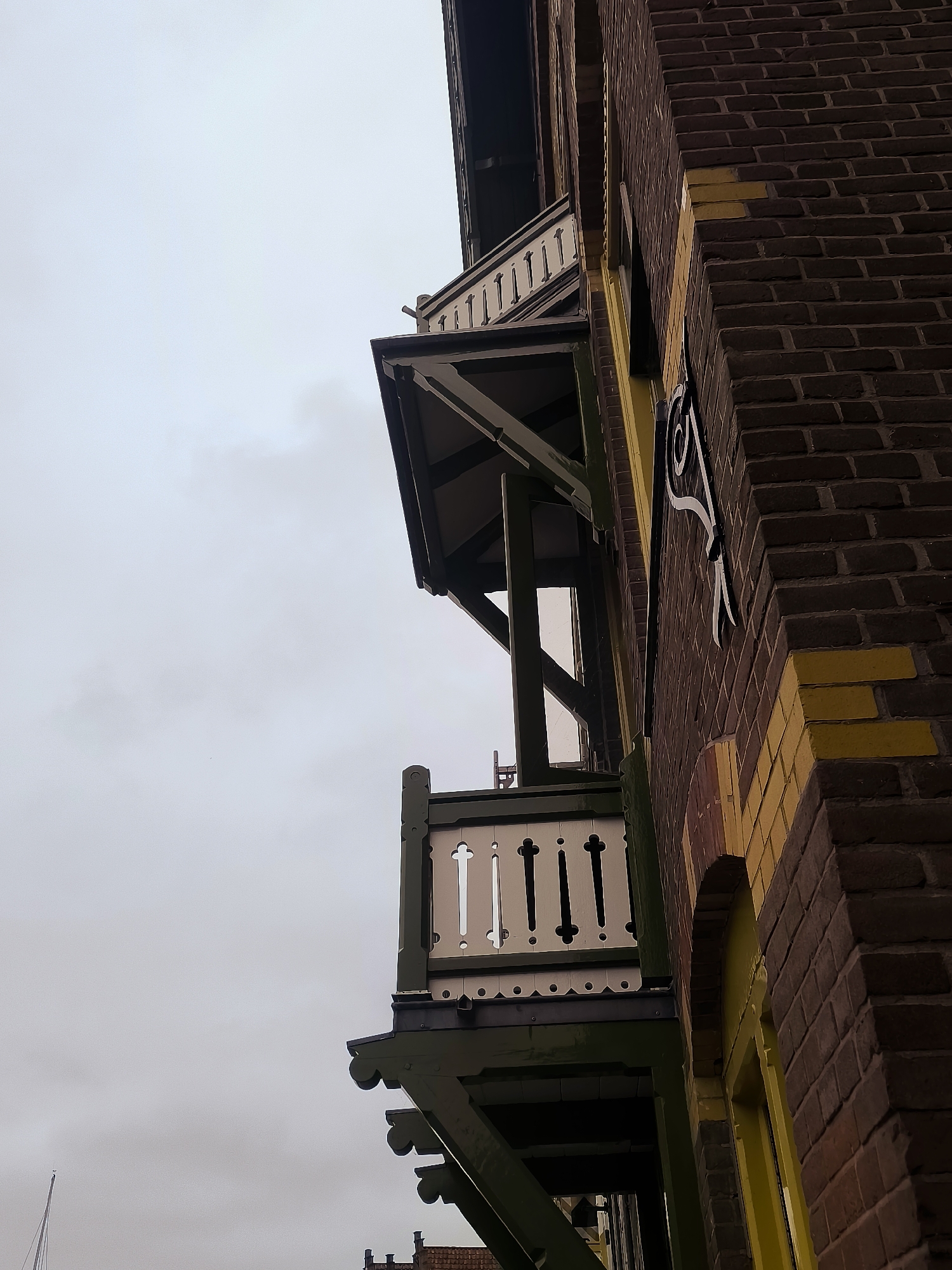 This is a low-angle, outdoor, medium shot of the side of a brick building in Hoorn, Netherlands. The building has a dark brick facade with yellow brick accents. There are two balconies visible, one above the other. The lower balcony has a white decorative railing and is supported by dark green wooden beams. The upper balcony has a similar white railing and is covered by a dark green awning. The sky is overcast and light gray. The overall impression is one of an older, possibly historic building.