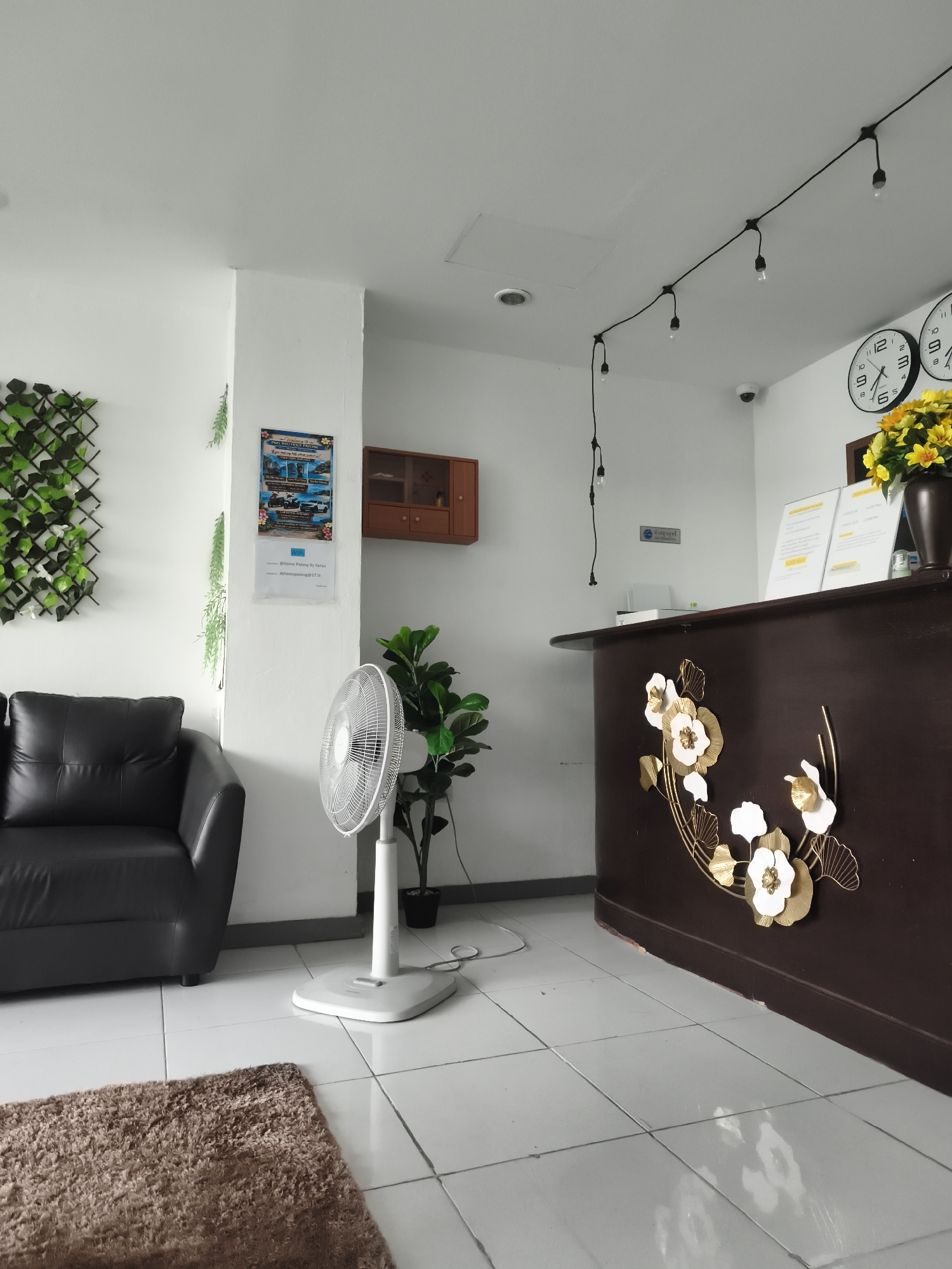 The image depicts the reception area of a hotel in Patong, Thailand. The scene is indoors, featuring a dark brown reception desk adorned with gold and white floral embellishments. Behind the desk, two clocks are mounted on the wall, displaying different times. A vase of yellow flowers sits on the counter, next to some documents. Overhead, a string of lights is suspended from the ceiling.
To the left of the desk, a black leather sofa is positioned near a wall with a decorative green plant lattice. In front of the sofa, a white oscillating fan stands on the tiled floor, beside a tall potted plant. A small brown rug is placed on the floor in the foreground. A framed poster with images and text, likely promotional material, is affixed to the wall near the sofa. A small wooden cabinet is mounted on the wall further left. A security camera is visible on the ceiling. The floor is tiled with light-colored squares. The overall environment suggests a welcoming and functional reception space.