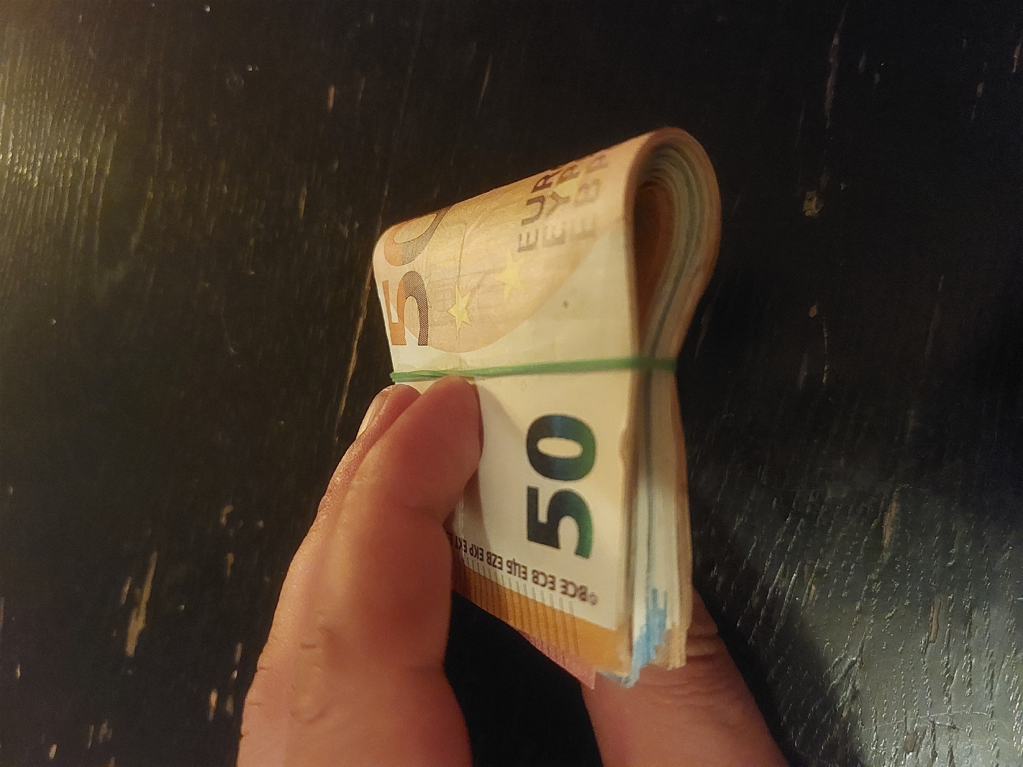 A hand holds a thick stack of 50 Euro banknotes, bound together by a green rubber band, against a dark, worn wooden surface. The location is not discernible from the image.