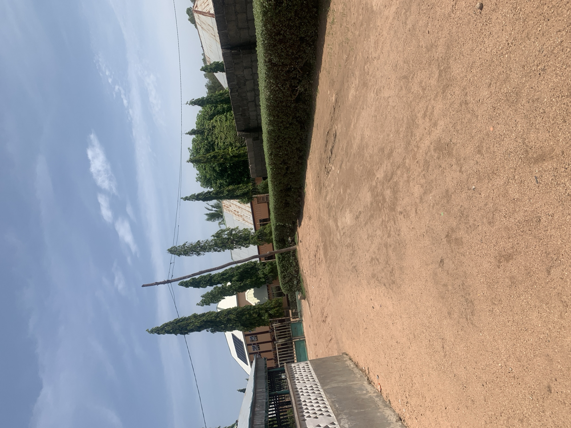 The image depicts an outdoor scene in Jauro Yinu, Nigeria, featuring a dirt pathway in the foreground. To the left, a gray wall with a decorative pattern runs along the edge of the pathway. Beyond the wall, a covered structure with a metal roof and supporting posts is visible. Further into the scene, a green metal gate is situated. Behind the gate, a building with a light-colored facade and solar panels on its roof is present. A line of tall, slender trees stands between the buildings and the pathway. To the right, a dense green hedge lines the pathway, followed by a gray brick wall and more buildings with corrugated metal roofs. The sky above is blue with scattered white clouds. The ground is a reddish-brown dirt, appearing dry and slightly uneven. The overall environment suggests a residential or institutional setting.