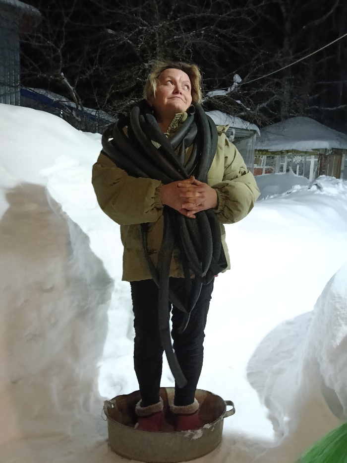 A woman stands outside on a snowy night. She is wearing a tan jacket, black pants, and red slippers, and she has dark tubes wrapped around her neck and bundled in her arms. She is standing in a metal wash tub, surrounded by deep snow. In the background, there are buildings covered in snow, as well as trees. The woman is looking upward with a contemplative expression.