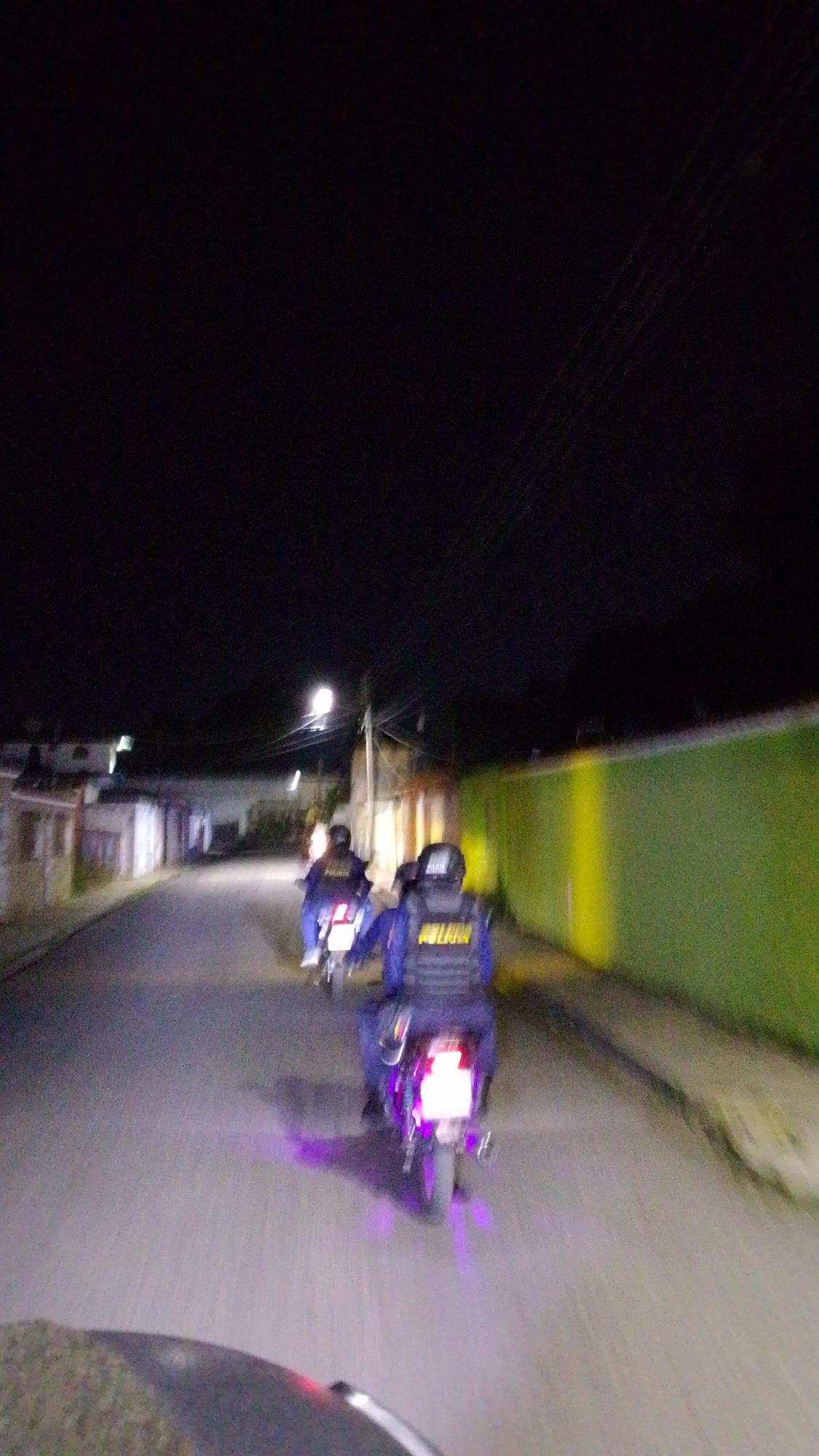 Two motorcyclists, possibly law enforcement officers ("POLICIA" visible on the vest of the closest rider), are traveling down a paved road at night. Streetlights illuminate the road ahead. Buildings are visible on the left side of the road, and a long green wall runs along the right. The scene appears to be in a residential area. The vehicles are moving, indicated by the motion blur. The photo was taken from the perspective of someone in a vehicle following the motorcyclists. The location is Tinaquillo, Venezuela.