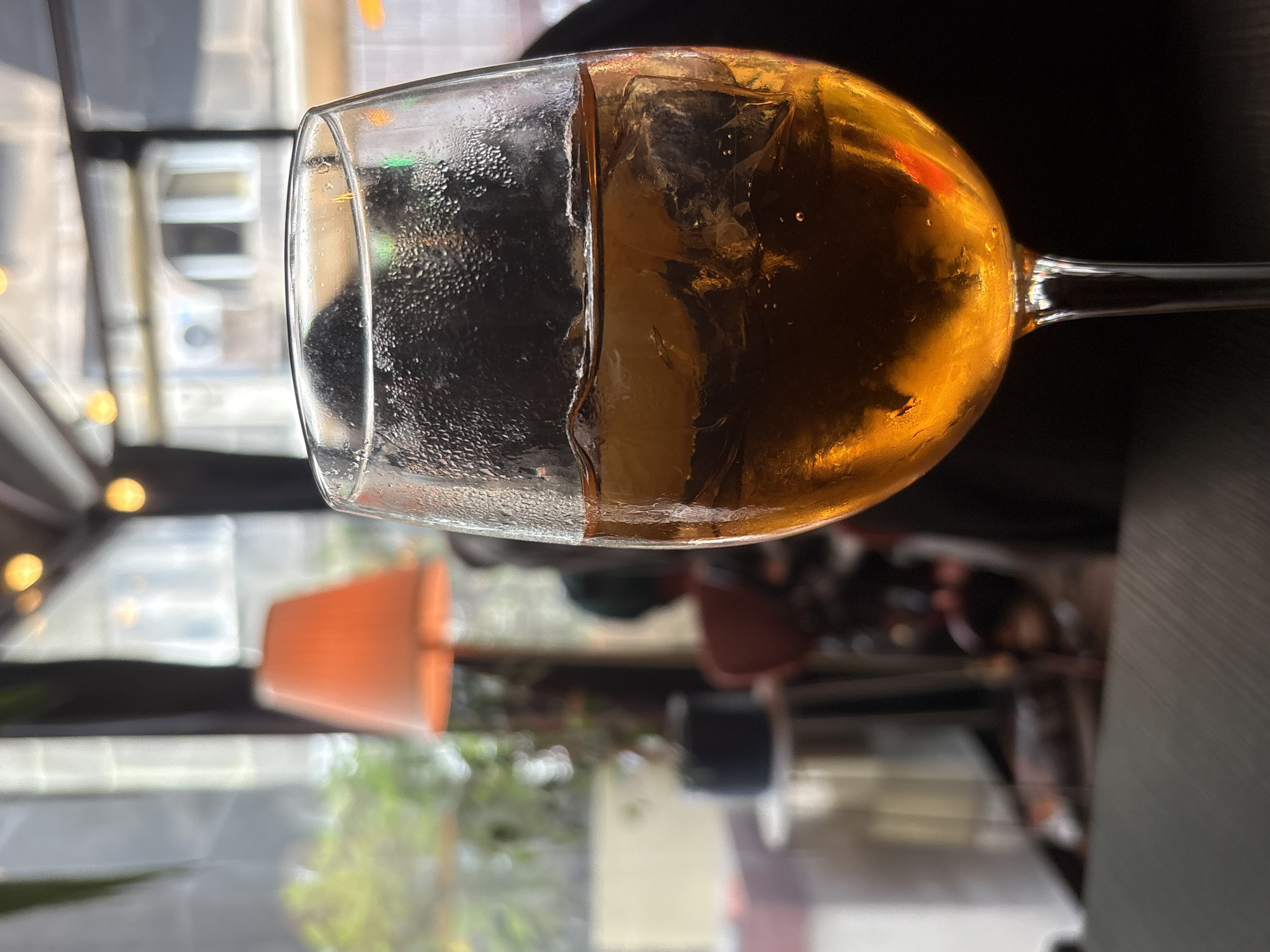 A wine glass, mostly filled with a golden-brown liquid, likely wine or a cocktail, sits on a reflective surface. The glass is noticeably cold, indicated by the condensation on its outer surface. Inside the glass, ice cubes are visible.
The background, visible through the glass and reflected in the surface beneath it, suggests an urban setting. Out of focus, we see elements of a city. The reflection shows a partial view of Belgrade, Serbia.