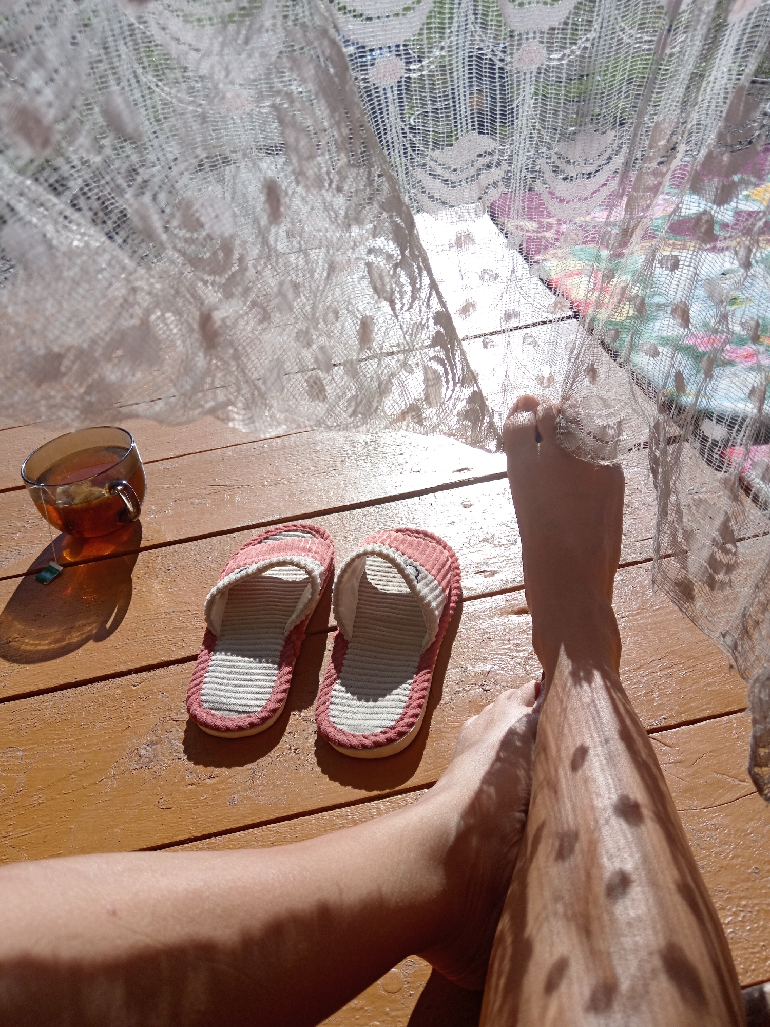 The image depicts a close-up, high-angle view of a person's legs and feet resting on a wooden porch. Sunlight casts dappled shadows from a lace curtain onto the wooden surface and the person's skin. A glass mug filled with tea and a tea bag is placed to the left of the feet, and a pair of pink and white striped slippers are situated in front of the feet. The background is blurred, showing the lace curtain and what appears to be foliage and possibly colorful fabric behind it. The scene is set outdoors on a sunny day.