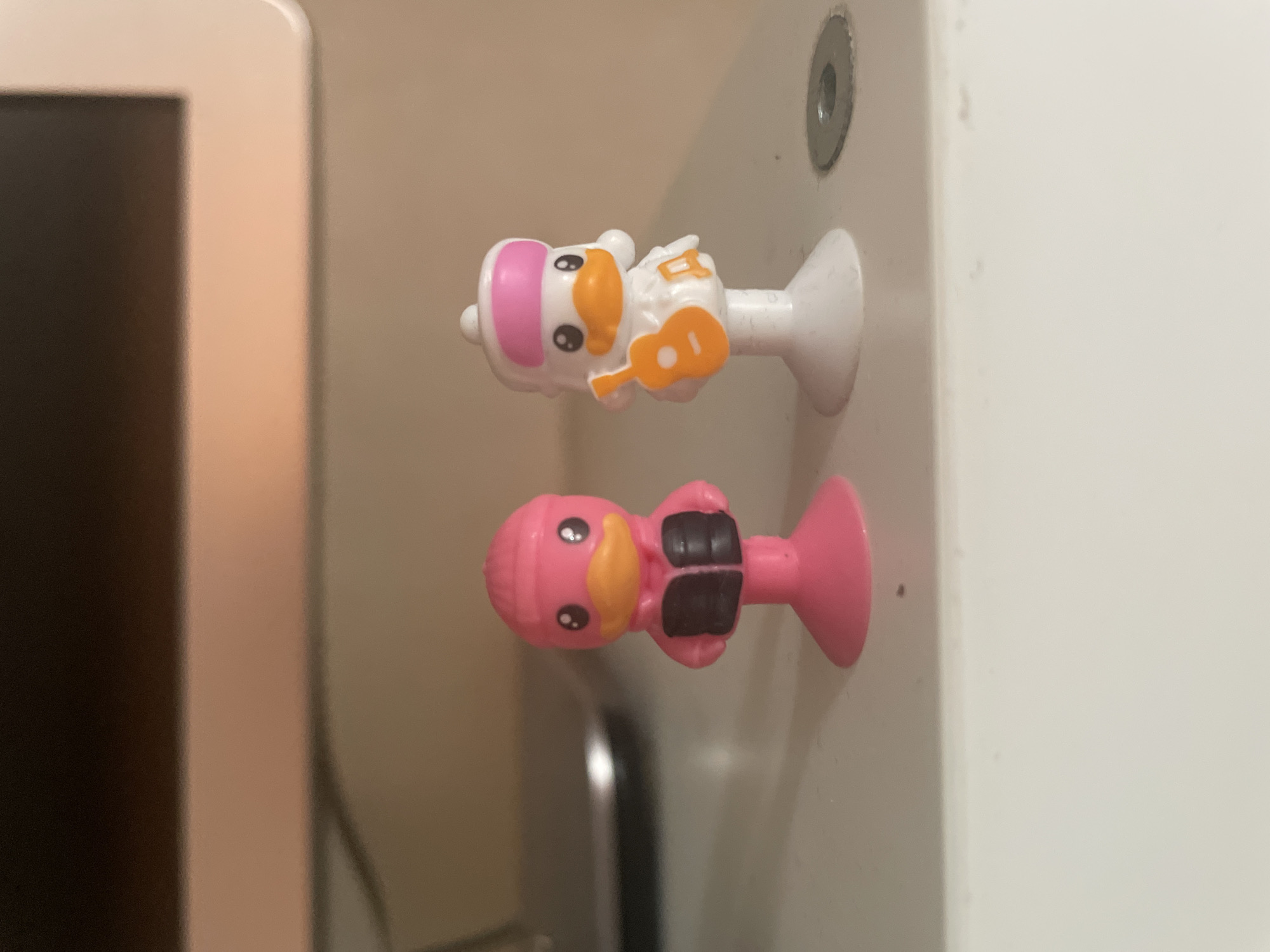 The image features two small, colorful plastic figures with suction cup bases, attached vertically to a light-colored surface. These figures are stylized cartoon ducks.
The upper figure is primarily white with a pink head covering, an orange beak, and black eyes with white highlights. It appears to be wearing a white textured outfit and holds a small orange guitar. Its suction cup is white.
The lower figure is entirely pink, also featuring an orange beak and black eyes with white highlights. It has a dark, possibly black, vest or harness-like detail on its body. Its suction cup is pink.
To the left of the figures, a portion of a light-colored electronic device, possibly a laptop or tablet, is visible, showing a dark screen and a light-colored bezel. The figures are mounted on a plain, light-colored surface, which could be a wall or a piece of furniture. Above the top figure, a metallic, circular object, resembling a screw head or a fixture, is embedded in this surface.
The scene appears to be indoors, under artificial lighting. The figures are stationary and no specific activity is occurring. There is no visible text in the image. The general atmosphere is that of a personal or domestic setting, adorned with these decorative items. No time of day, weather conditions, or specific emotions are discernible beyond the cute expressions on the figures.