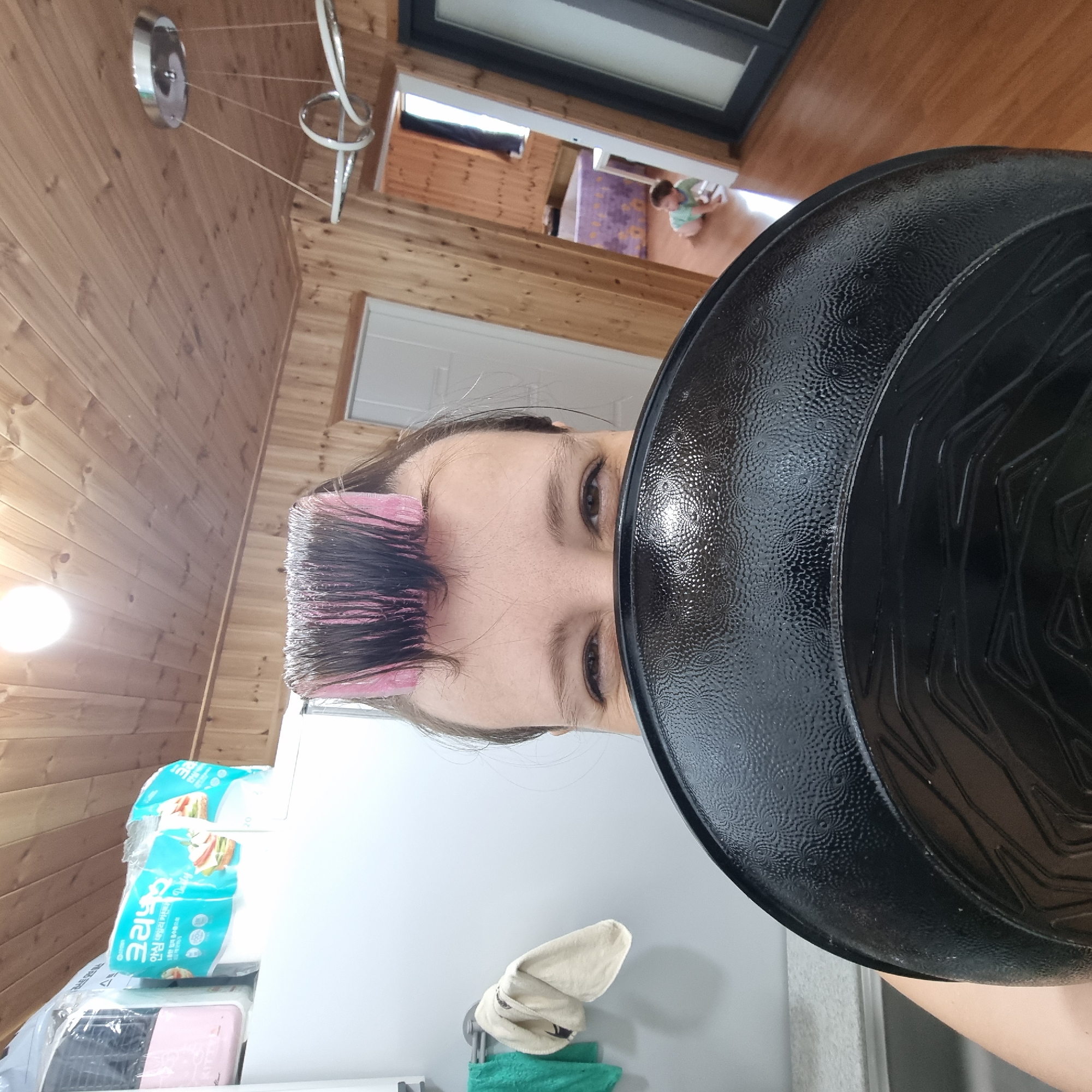 In a residential interior that appears to be in Dunnae-myeon, South Korea, a woman with pink hair rollers on her head is holding a black frying pan up to her face, obscuring most of her face. Only her eyes and forehead are visible. The background shows wooden paneled walls and ceiling, a chandelier, and a doorway leading to another room where a young child is crawling on the floor. To the left of the woman, there are towels hanging on a rack. Several packages of what appear to be facial tissues are stacked on a white surface. The lighting suggests it is daytime.