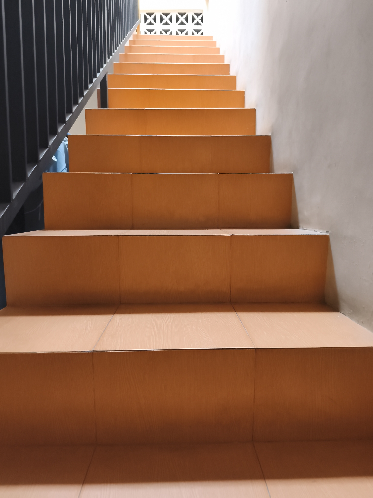 An upward view of an empty, orange-tiled staircase. The staircase ascends from the bottom of the frame to the top. A black metal railing runs along the left side of the stairs. The wall on the right side of the stairs is a light beige color. At the top of the stairs, a white decorative vent is visible, and beyond that, bright light suggests an exit or open space. The scene appears to be an interior or covered outdoor staircase, possibly in a building in Yogyakarta, Indonesia, given the architectural style and common building materials found in the region. There are no people or animals visible in the image.