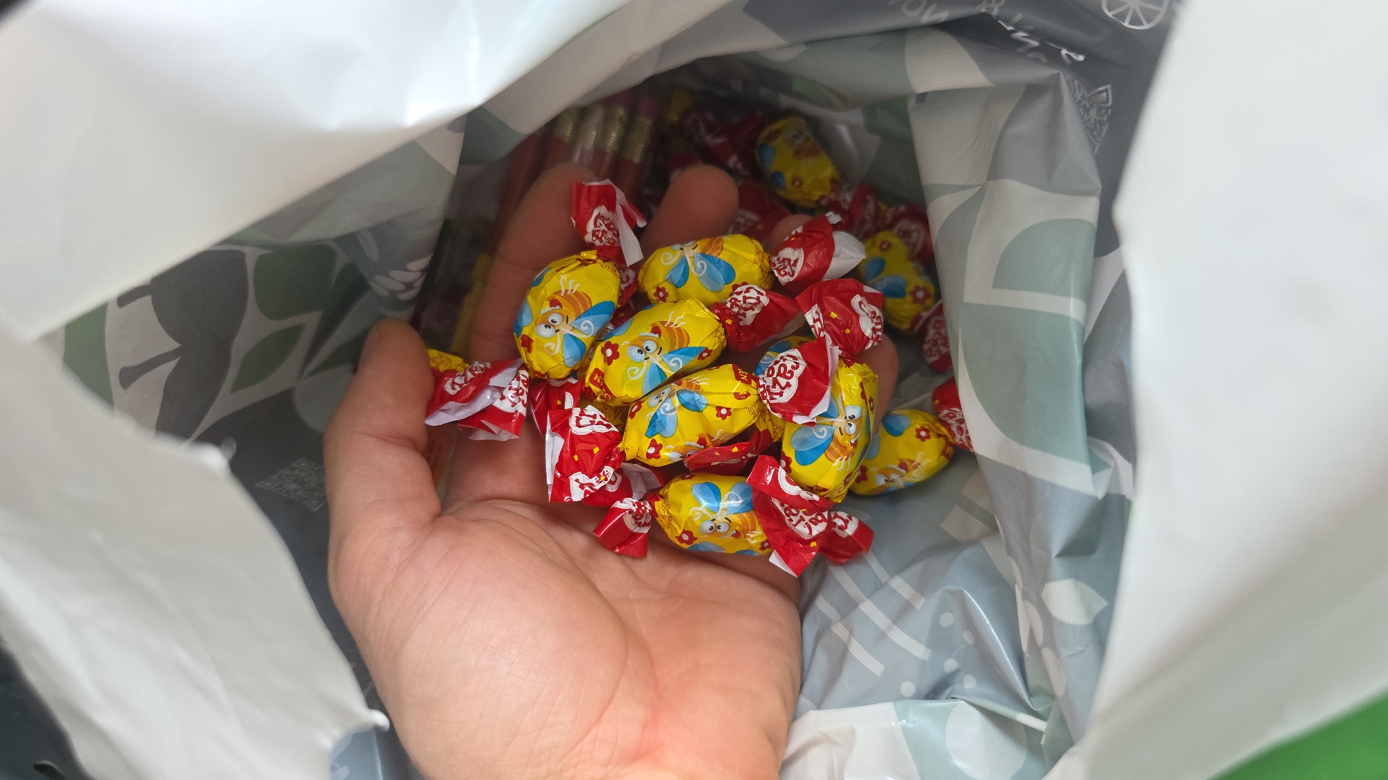 A close-up shot from above shows a person's hand holding a pile of individually wrapped candies. The candies are oval-shaped and feature a yellow wrapper with a cartoon bee and red flowers. Some of the wrappers are red with white writing. The candies are inside a white bag with a gray and light blue patterned lining. The bag appears to be a reusable shopping bag. The image is taken from a high angle, looking down into the bag. The location is not specified in the image or any provided context.