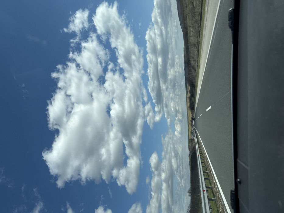 The image captures a forward-facing view from inside a vehicle traveling on a multi-lane highway near Nevsha, Bulgaria, under a bright blue sky dotted with numerous white cumulus clouds. The dark dashboard of the vehicle occupies the bottom of the frame. The asphalt road features clear white lane markings and extends into the distance. To the immediate left, a silver metal guardrail with white posts and small red reflectors lines the road, separated by a narrow grassy strip. To the right, a wide paved shoulder transitions to a grassy verge and then dry, undulating terrain with sparse, light brown vegetation. A rudimentary fence line with posts is visible on the right side of the landscape. Several dark-colored vehicles are discernible in the distance on the highway ahead. No individuals are visible. The scene suggests fair weather conditions during daylight hours.