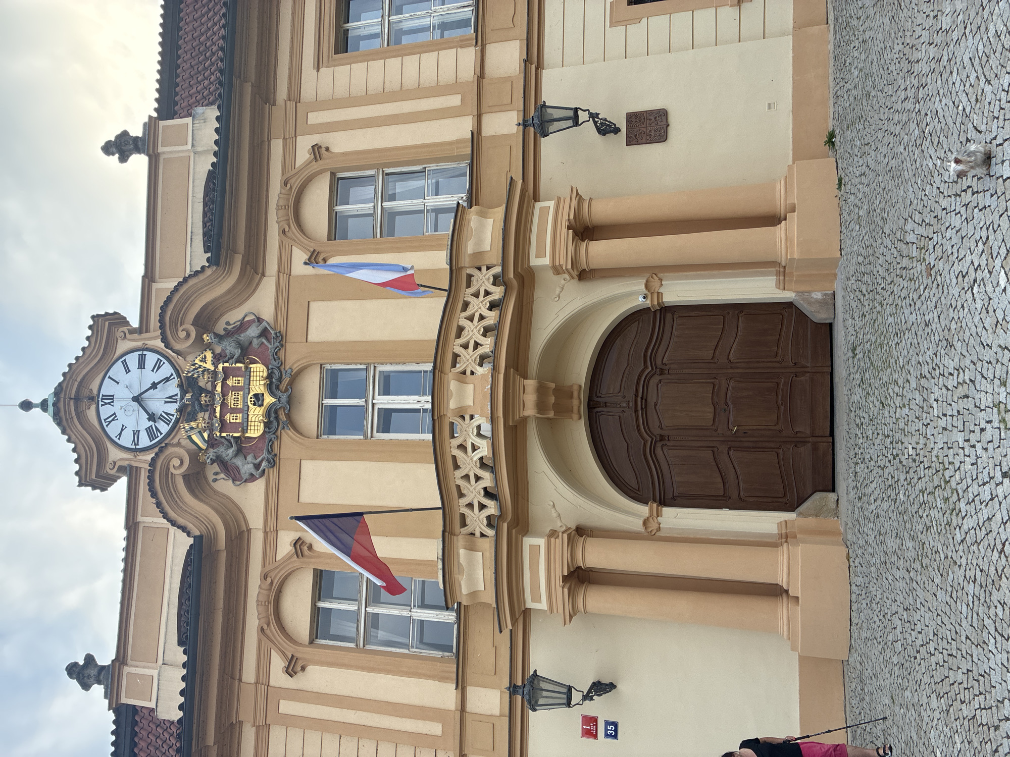 This image shows the exterior of the Archbishop's Palace in Prague, Czechia. The palace, with its distinct yellow facade and ornate architectural details, features a large, arched wooden entranceway flanked by two substantial columns. Above the entrance, a balcony with decorative railings extends across the building, adorned with two national flags, one of which appears to be the Czech flag. A large clock face is prominently displayed on the upper facade, positioned above a coat of arms featuring two lions supporting a shield. Decorative lamps are mounted on the walls on either side of the entrance. In front of the palace is a cobblestone plaza. To the left of the entrance, a woman with dark hair, wearing a black top and pink shorts, is standing with trekking poles. To the right of the entrance, near the bottom right corner of the image, a small, light-colored dog is visible on the cobblestones. A sign with the numbers "1" and "35" is attached to the wall to the left of the entrance. The sky overhead is overcast.