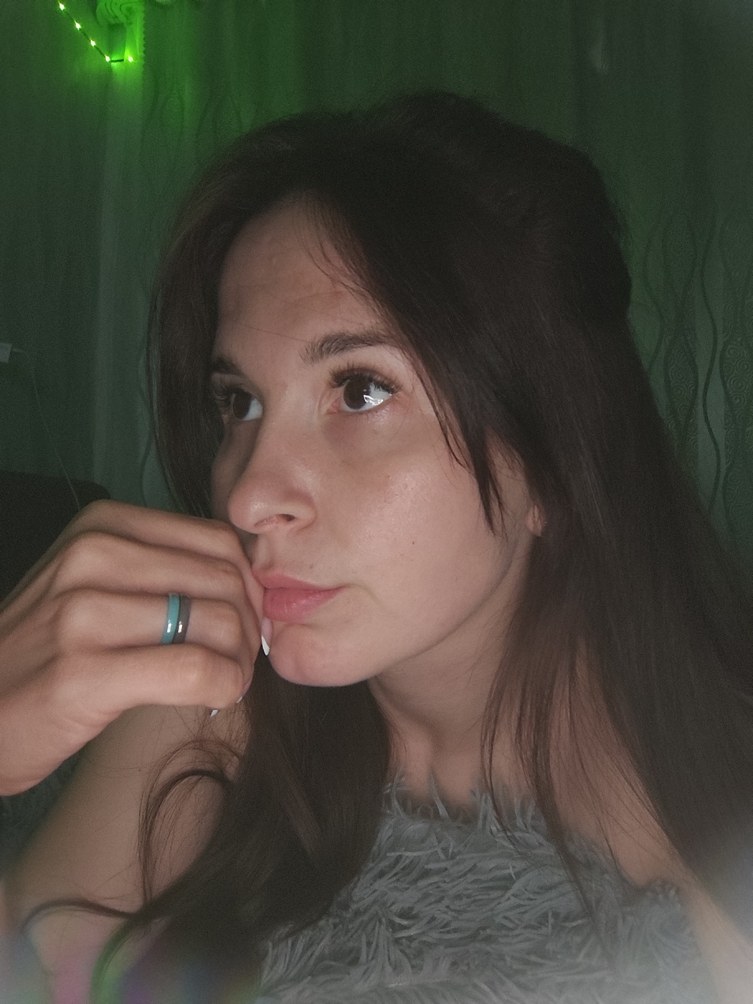 A medium close-up portrait features a young woman with long, dark brown hair, looking thoughtfully towards the viewer's left. Her left hand is raised, with her index finger gently touching her chin or lower lip, and her nails are manicured with white tips. She wears a unique two-toned ring on her left ring finger, featuring sections of teal/turquoise and a darker grey/silver. She appears to be wearing or covered by a soft, fluffy, light grey garment or blanket. Her expression is pensive and calm.
The setting is indoors, with a dimly lit background dominated by a green hue, which emanates from green string lights visible in the upper left corner. The wall behind her has a subtle patterned texture. The lighting suggests it is evening or nighttime. No text is visible in the image. The scene depicts a quiet, personal moment within a residence in Фоки, Russia.