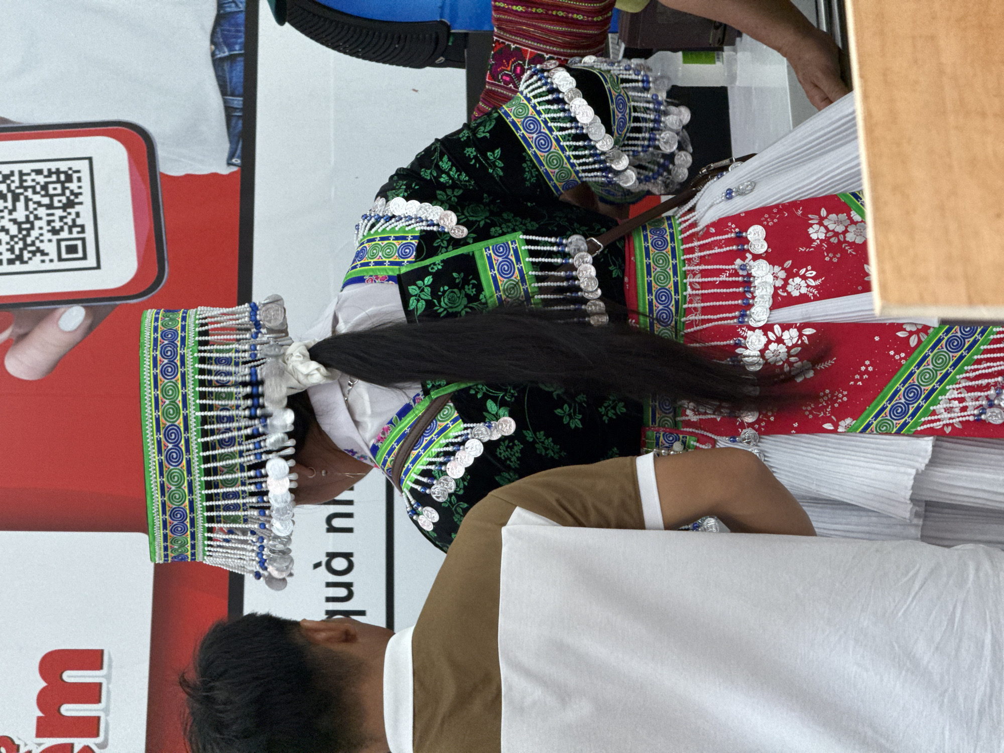 A person in traditional Hmong attire, featuring a black headdress adorned with silver coins and a green and blue patterned design, stands next to another person with short black hair, who is wearing a brown shirt and white apron. The environment includes promotional signage with a QR code and red lettering. The scene appears to be in a public setting or market environment, as the setting aligns with traditional Hmong locations.