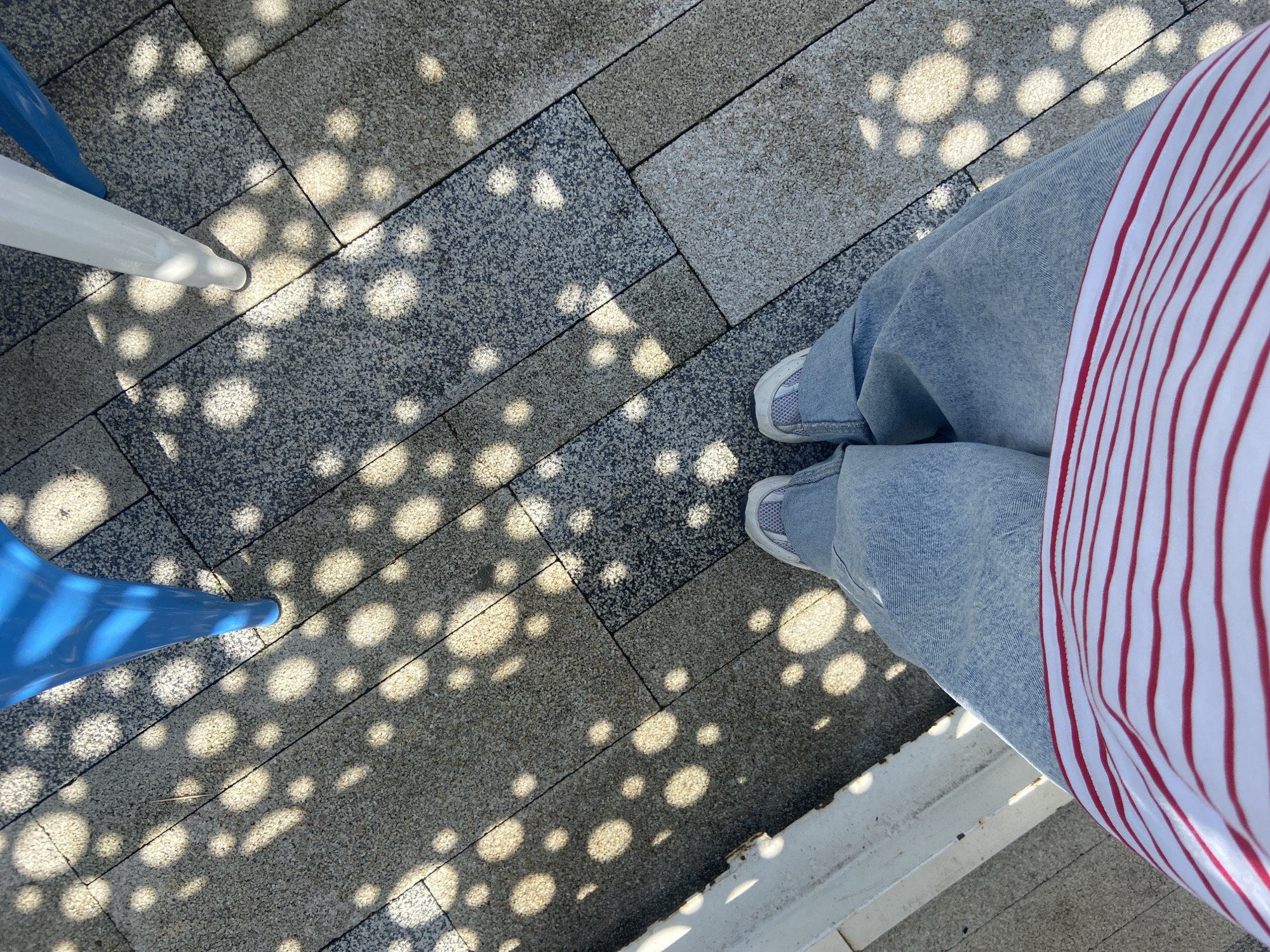The image shows a person standing on a brick patio with dappled sunlight filtering through an overhead structure, creating circular shadows. The person is wearing light-wash denim jeans, white sneakers, and a red and white striped shirt. The patio is made of grey and tan brick tiles. The legs of a blue and white chair are visible on the left. The image includes a view from above of the person's feet and legs. The location is not provided in the image.