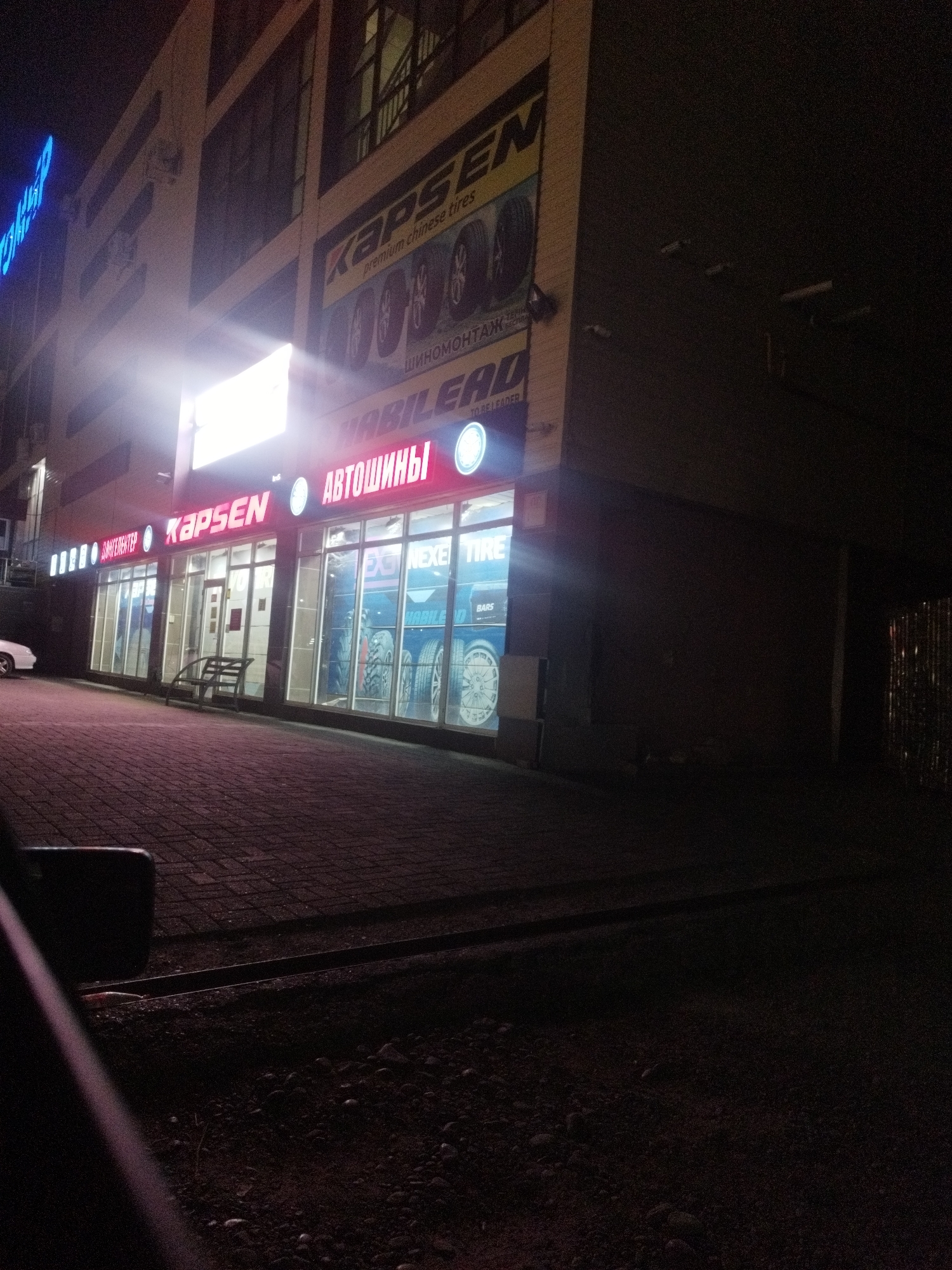 The image shows a tire shop named "KAPSEN" located on a street in Shymkent, Kazakhstan, at night. The shop's facade is illuminated by bright neon signs that read "KAPSEN" and "АВТОШИНЫ," which translates to "TIRES." Large glass windows display various tires and wheel rims. A white car is parked to the left of the entrance. The ground in front of the shop is paved with interlocking bricks, and a metal bench is placed near the entrance. The street appears to be a commercial area with other buildings visible in the background. The overall atmosphere is illuminated by artificial light sources.