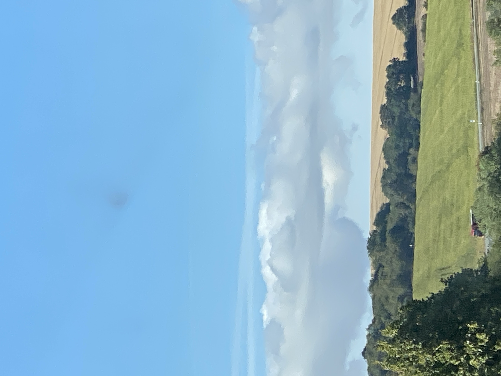 The image depicts a rural landscape under a bright blue sky with scattered white clouds. In the foreground, a lush green field slopes upwards, bordered by a treeline. Beyond the treeline, a field of harvested golden crops stretches towards the horizon. A red truck is visible on a road in the lower right portion of the image, and a few figures can be seen in the distance on the golden field. The overall impression is one of a serene, agricultural setting. Without further information or specific landmarks, it is impossible to pinpoint the exact location within Nordhausen, Germany.