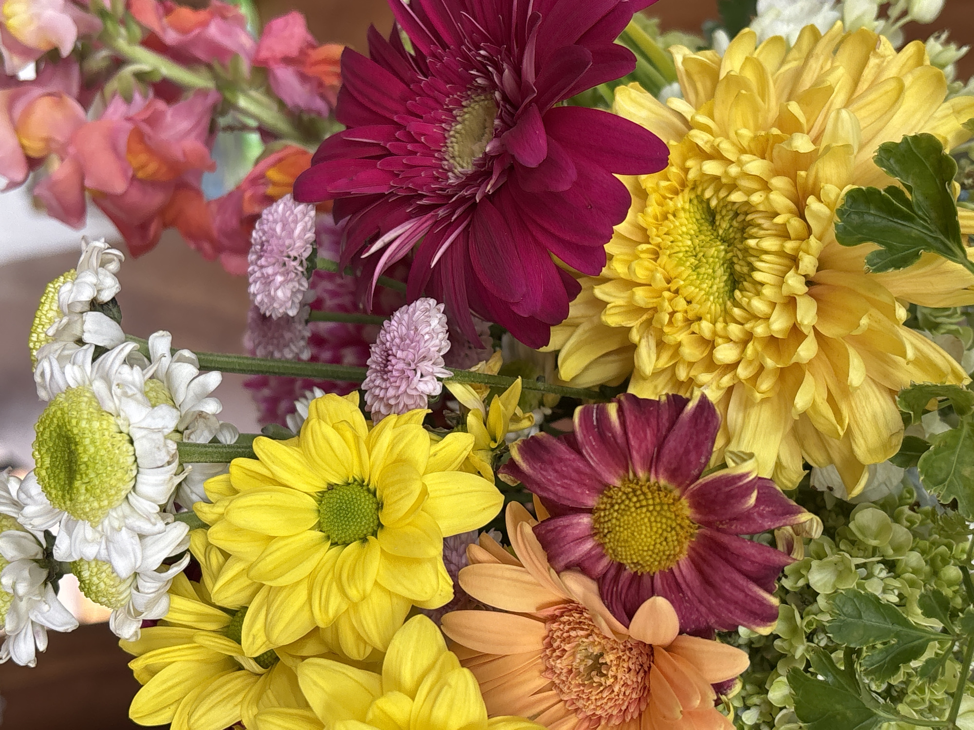 The image presents a close-up view of a bouquet of colorful flowers. Dominant colors include yellow, deep red, white, and orange. Several types of flowers are visible, including chrysanthemums (yellow and white), gerbera daisies (deep red and orange), and smaller, round pink flowers. There is also what appears to be snapdragons in the upper left corner and small green foliage. The background is slightly out of focus, providing a shallow depth of field. No people are visible. The location of the image is not identifiable from its contents.