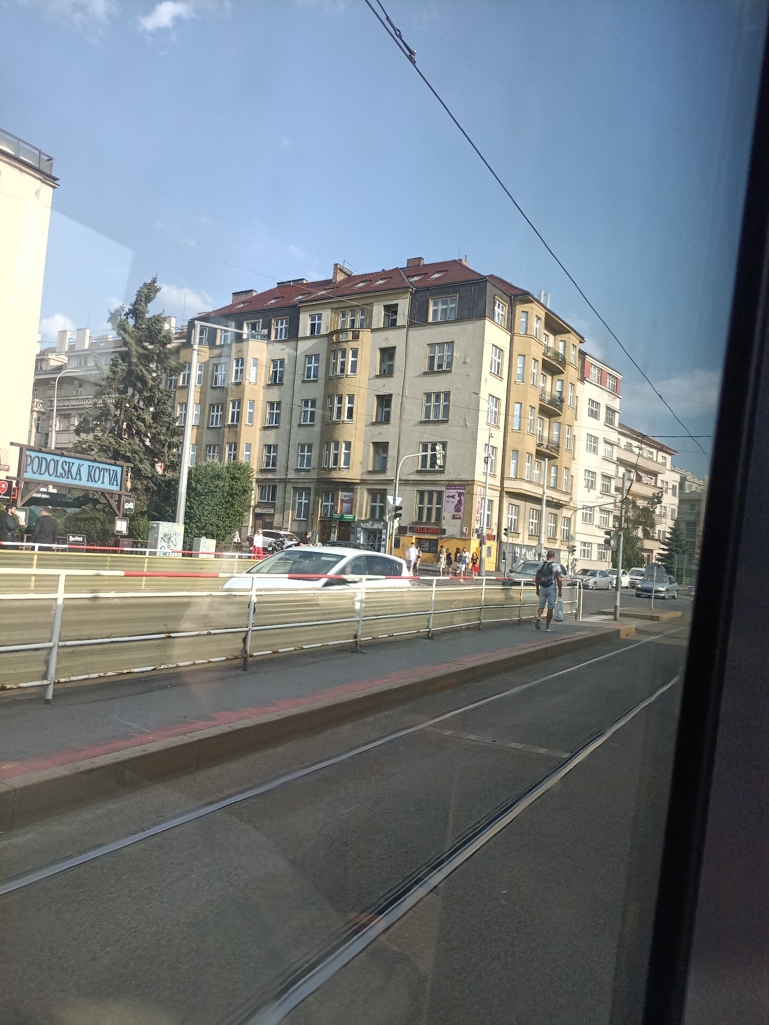 The image captures a view of a street in Prague, Czechia, from the perspective of a moving vehicle, likely a tram. The scene features an urban environment with multi-story buildings lining the street. A prominent sign with blue text reading "PODOLSKA KOTVA" is visible on the left side of the image, indicating a specific location or establishment. Several cars are present on the road, including a white car and other vehicles further down the street. Pedestrians are also visible on the sidewalk. The street is equipped with tram tracks, suggesting that this is a common route for public transportation. The architecture of the buildings is characteristic of European cities, with multiple windows and varied rooflines. The sky above is partly cloudy. The perspective suggests the image was taken during daylight hours.