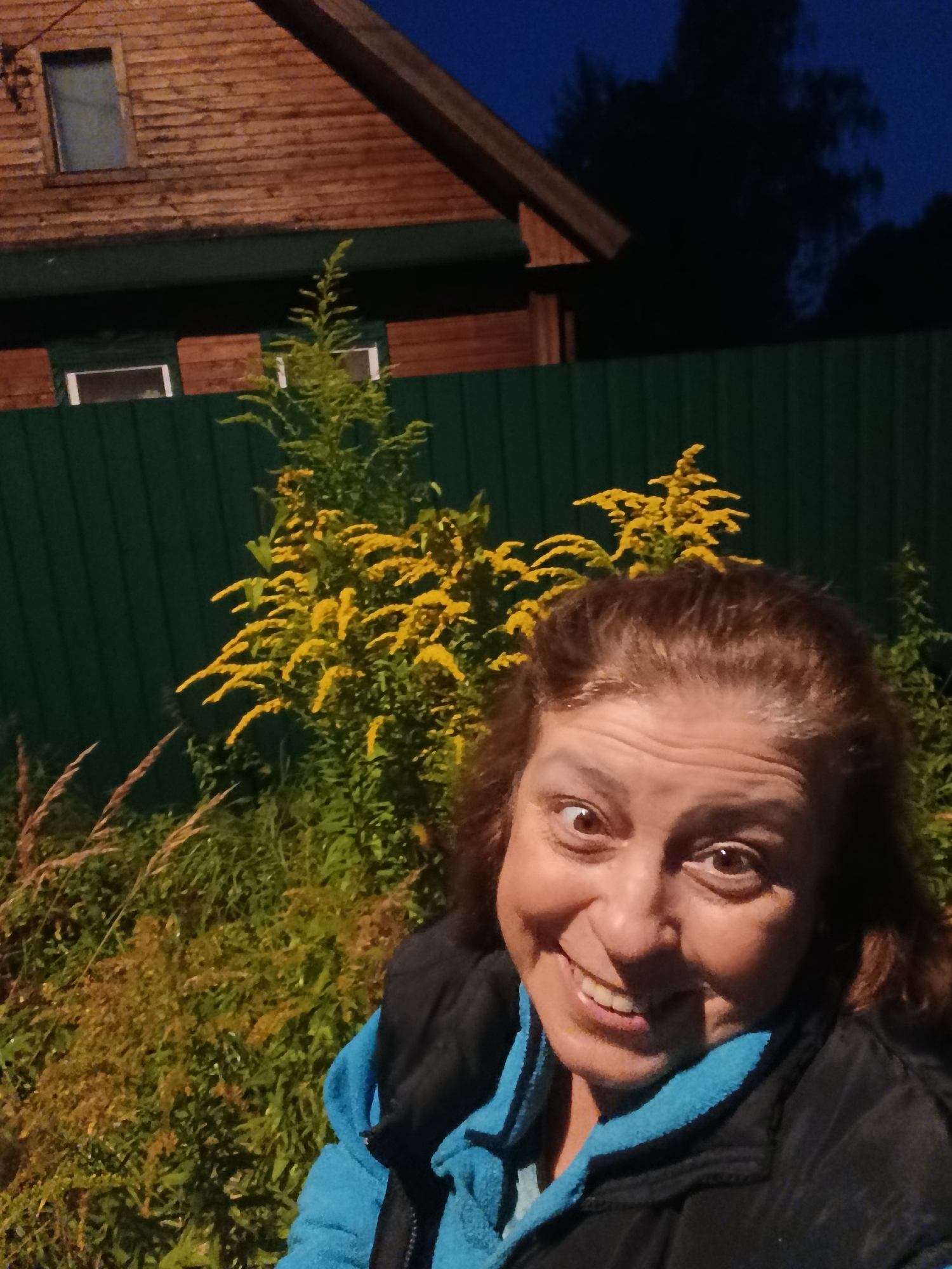 A selfie of a woman with brown hair and brown eyes is taken outdoors in front of a wooden house and a green fence. The woman is wearing a black vest over a blue hooded jacket. Behind her, bright yellow flowering plants are visible, along with tall grasses. The sky is dark blue, indicating it is evening or night. The wooden house has horizontal siding and a visible roof structure. The overall scene suggests a rural or suburban setting.