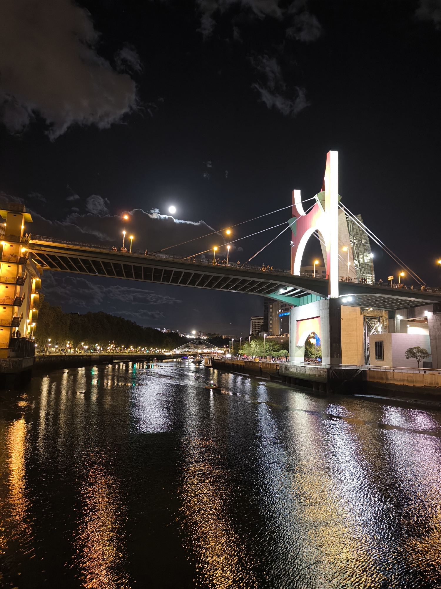 The image depicts the city of Bilbao, Spain, at night. A prominent suspension bridge with illuminated white and red arch structures dominates the right side of the frame. The bridge carries vehicular traffic and pedestrians, with visible figures walking across its deck. Below the bridge, a body of water, likely the Nervión River, reflects the city lights, creating shimmering patterns on its surface. On the left bank, a building with warm yellow lights and balconies is visible, adjacent to trees and a promenade lined with streetlights. Further in the distance, other bridges and city buildings are illuminated against the dark night sky. A full moon is visible in the upper center of the sky, partially obscured by clouds. Several small boats are present on the water. The overall scene captures the vibrant atmosphere of Bilbao at night, highlighting its architectural landmarks and waterfront.