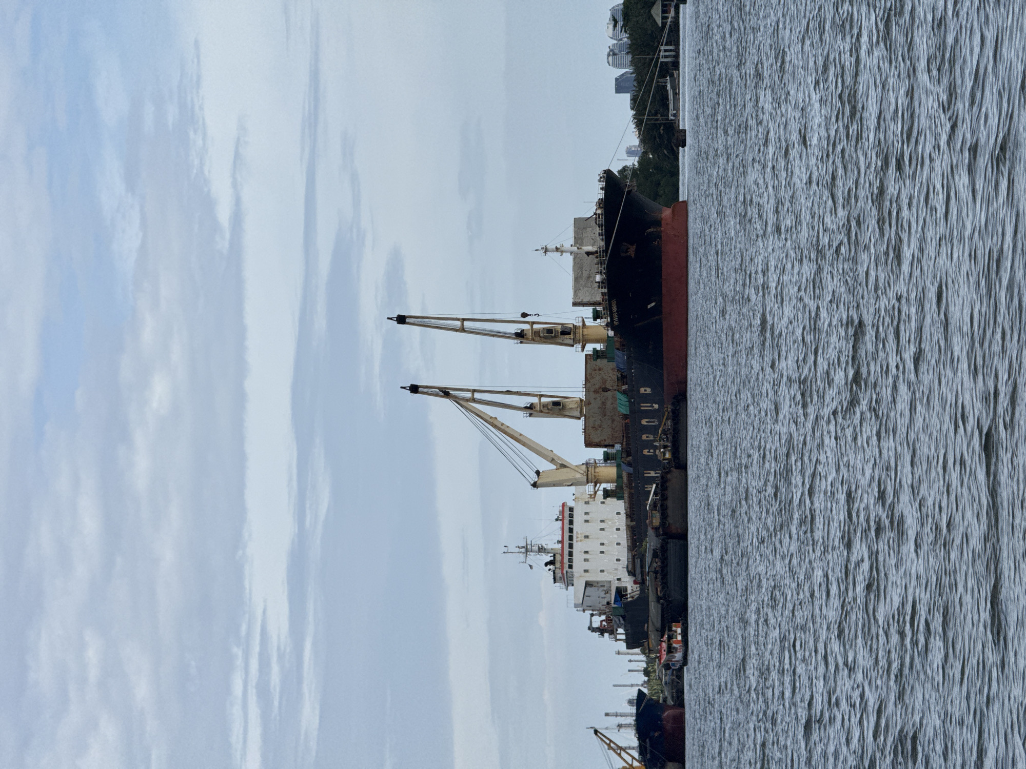 The image depicts a wide river in Bangkok, Thailand, with several large cargo ships docked along its banks. The sky is overcast with a light blue hue and scattered clouds. The foreground is dominated by the choppy surface of the river, reflecting the muted light.
In the midground, the primary focus is a substantial cargo ship with a white superstructure and a red hull. This vessel is equipped with two large cranes, one extending to the left and the other to the right. The ship's name, "H GROUP," is visible on its side. Alongside this main ship, there are several barges and smaller vessels. To the left, another large blue-hulled ship is partially visible, also with a crane. Further in the background, industrial structures and the silhouette of buildings are discernible against the horizon, suggesting an urban or industrial area within Bangkok. The overall scene conveys a sense of industrial activity and port operations. There are no visible people in the frame.