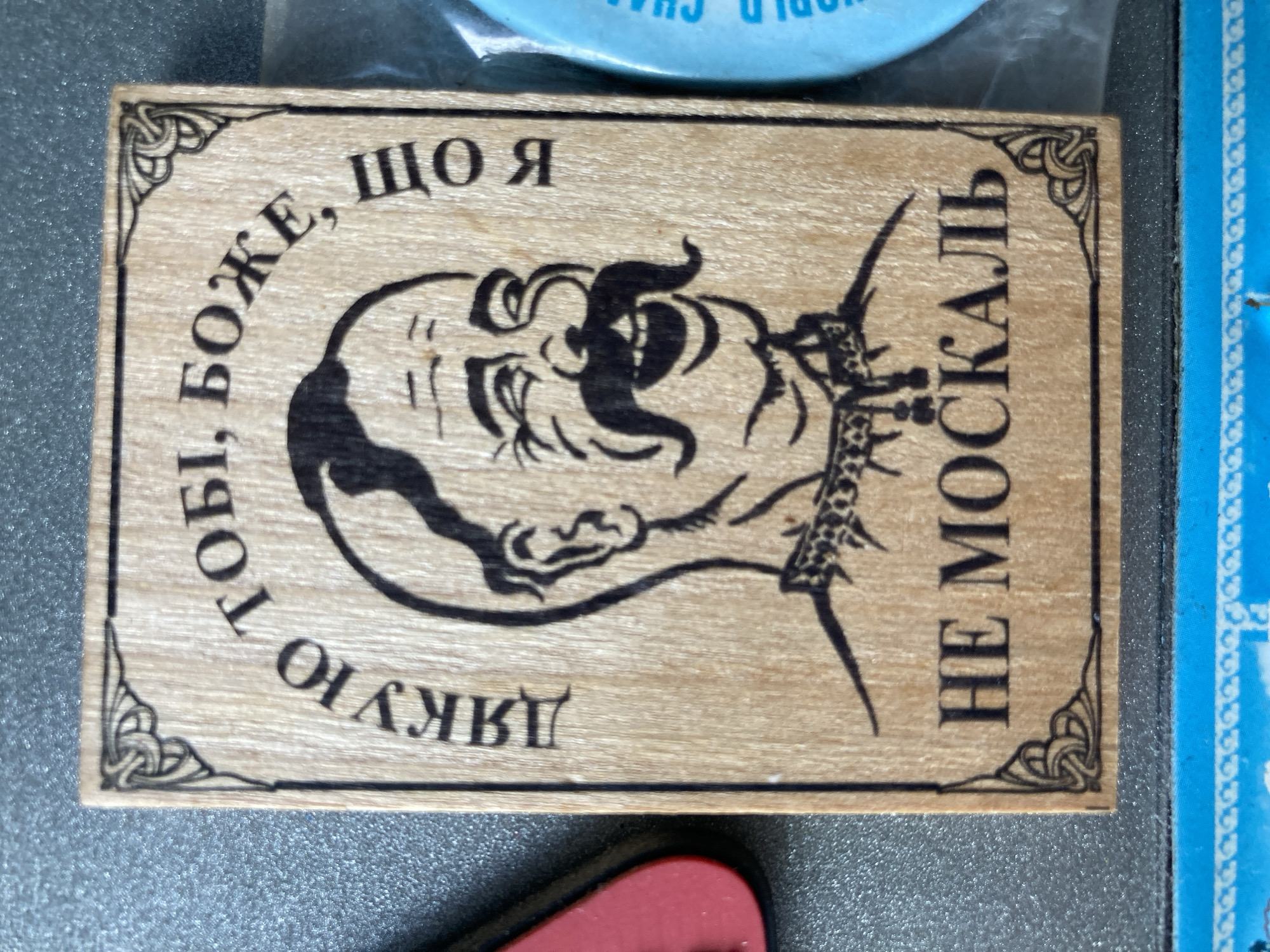 A wooden souvenir magnet is displayed on a textured grey surface. The magnet is rectangular with rounded corners and features a black illustration of a man's head with a large mustache and a wide smile, appearing to be laughing. Above the man's head, curved text in Russian reads, "ДЯКУЮ ТОБІ, БОЖЕ, ЩО Я НЕ МОСКАЛЬ" which translates to "Thank you, God, that I am not a Moskal". The word "МОСКАЛЬ" (Moskal) is a derogatory term for Russians. The magnet also has decorative Celtic knot designs in the corners. To the left of the magnet, a portion of a red heart-shaped object is visible. To the right, a blue object with white text, part of which reads "WORLD CHA", is also partially visible. The overall presentation suggests the magnet is attached to a metallic surface, possibly a refrigerator or a locker.