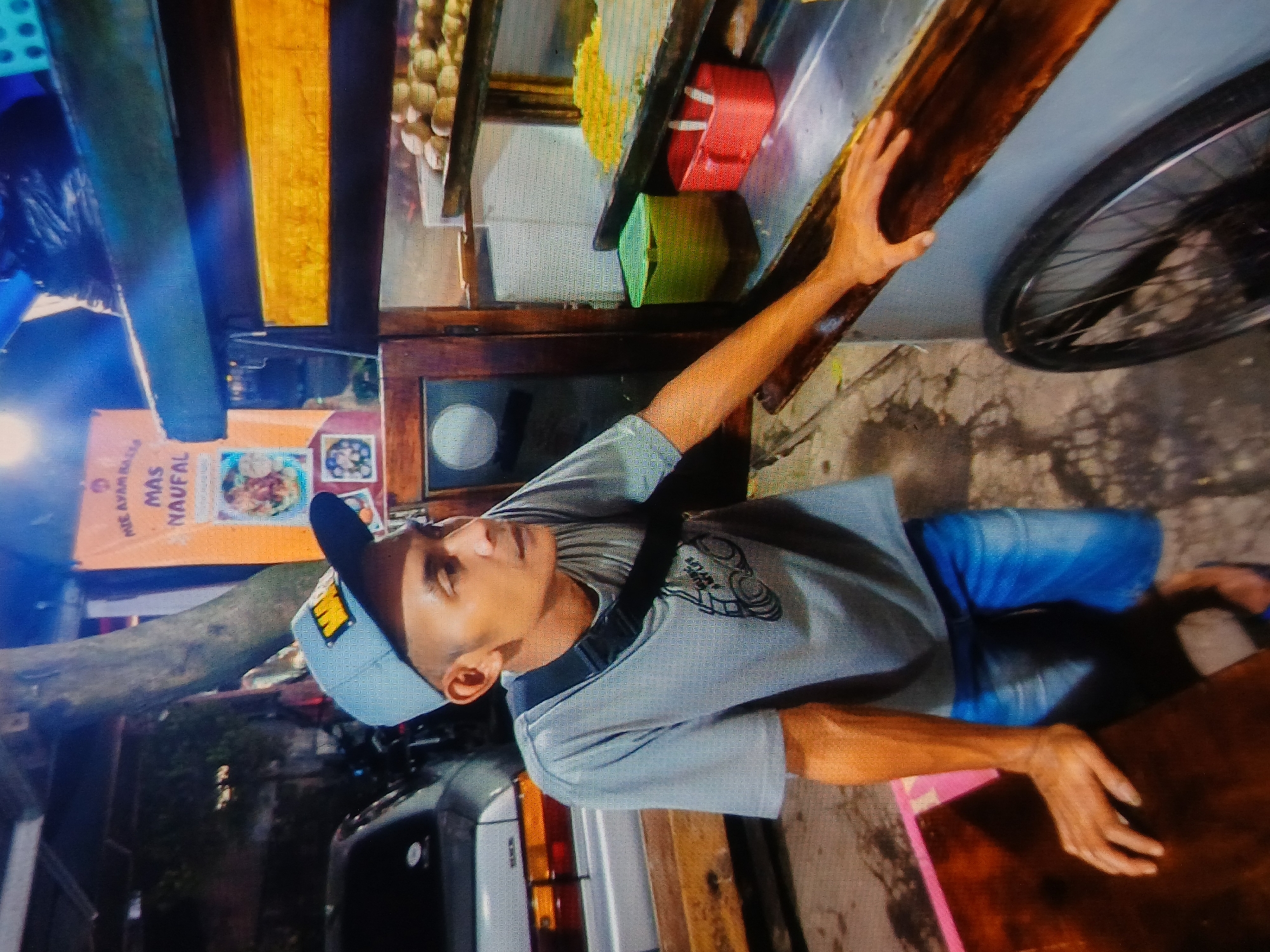 A medium shot captures a man wearing a blue baseball cap and a gray t-shirt with a graphic design, standing next to a food cart. The man is positioned in the foreground, with his left arm resting on a wooden table and his right hand leaning on the food cart. He appears to be looking to his right, with his mouth slightly open. The food cart, likely located in Setia Bakti, Indonesia, is filled with various food items and containers. A large orange banner with text in Indonesian hangs in the background, above the cart. A silver car is partially visible to the left of the frame. The scene is lit by artificial light, creating a warm glow and deep shadows. The overall impression is that of an outdoor food stall at night.