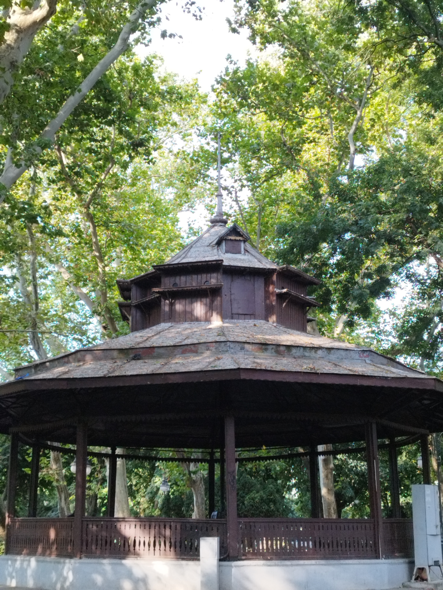 The image captures a hexagonal wooden gazebo situated within a park. The gazebo is dark brown and features a multi-layered roof. The lower part of the structure is enclosed by a decorative wooden railing. Tall trees with lush green foliage surround the gazebo, creating a shaded environment. The sky is visible through the canopy of the trees. The gazebo appears to be located in Bucharest, Romania, possibly in a public park or garden. There are no people clearly visible in the image. The lighting suggests it is daytime, with sunlight filtering through the leaves.
