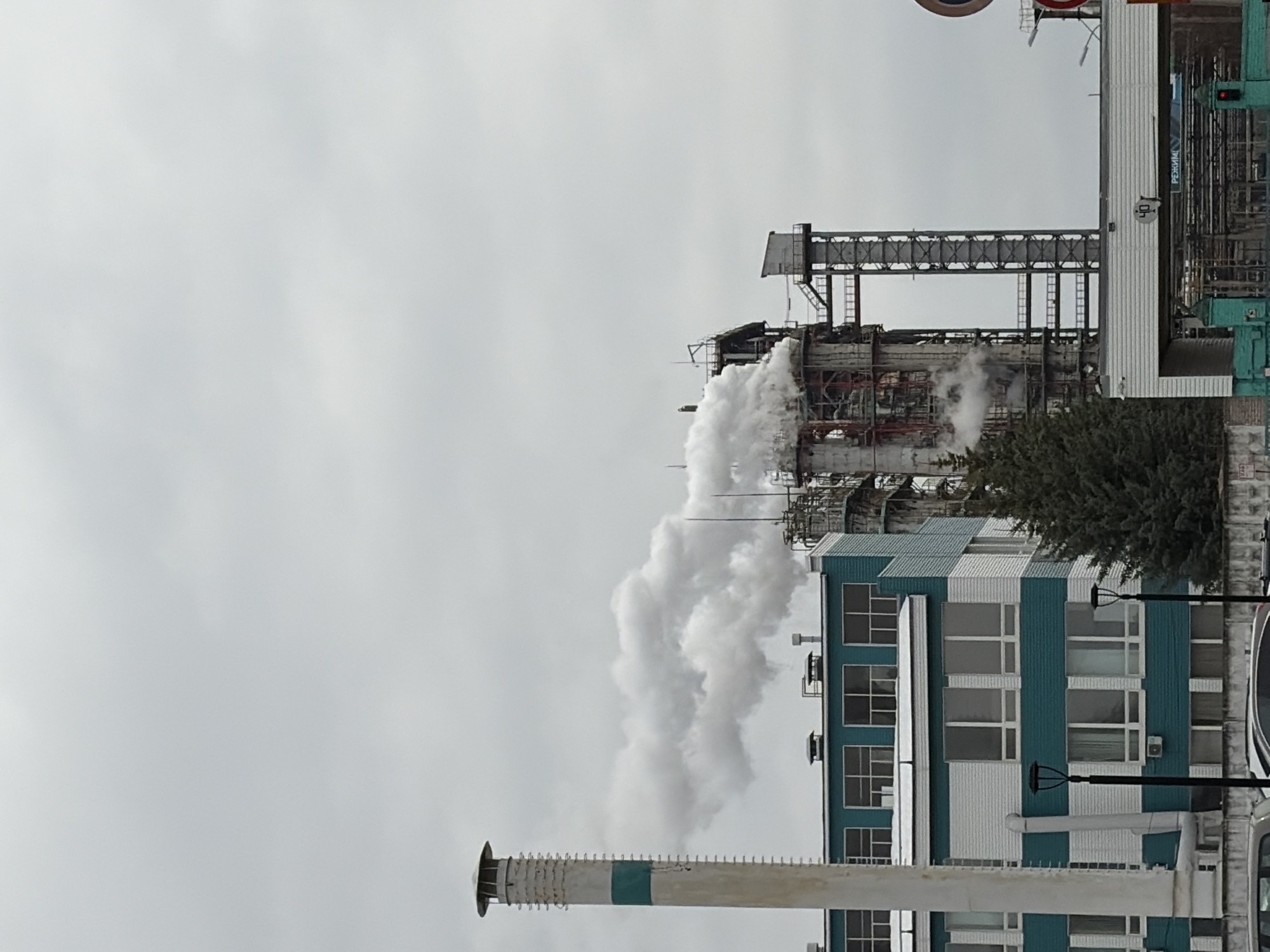 The image shows an industrial landscape in Chernushka, Russia. In the foreground, a tall chimney with a white and teal striped pattern stretches towards the sky. The background features an industrial complex, which consists of a multi-storied turquoise and white building with rows of windows and a steel structure rising above it. Dense white smoke is emanating from the steel structure. The sky is overcast and grey.
