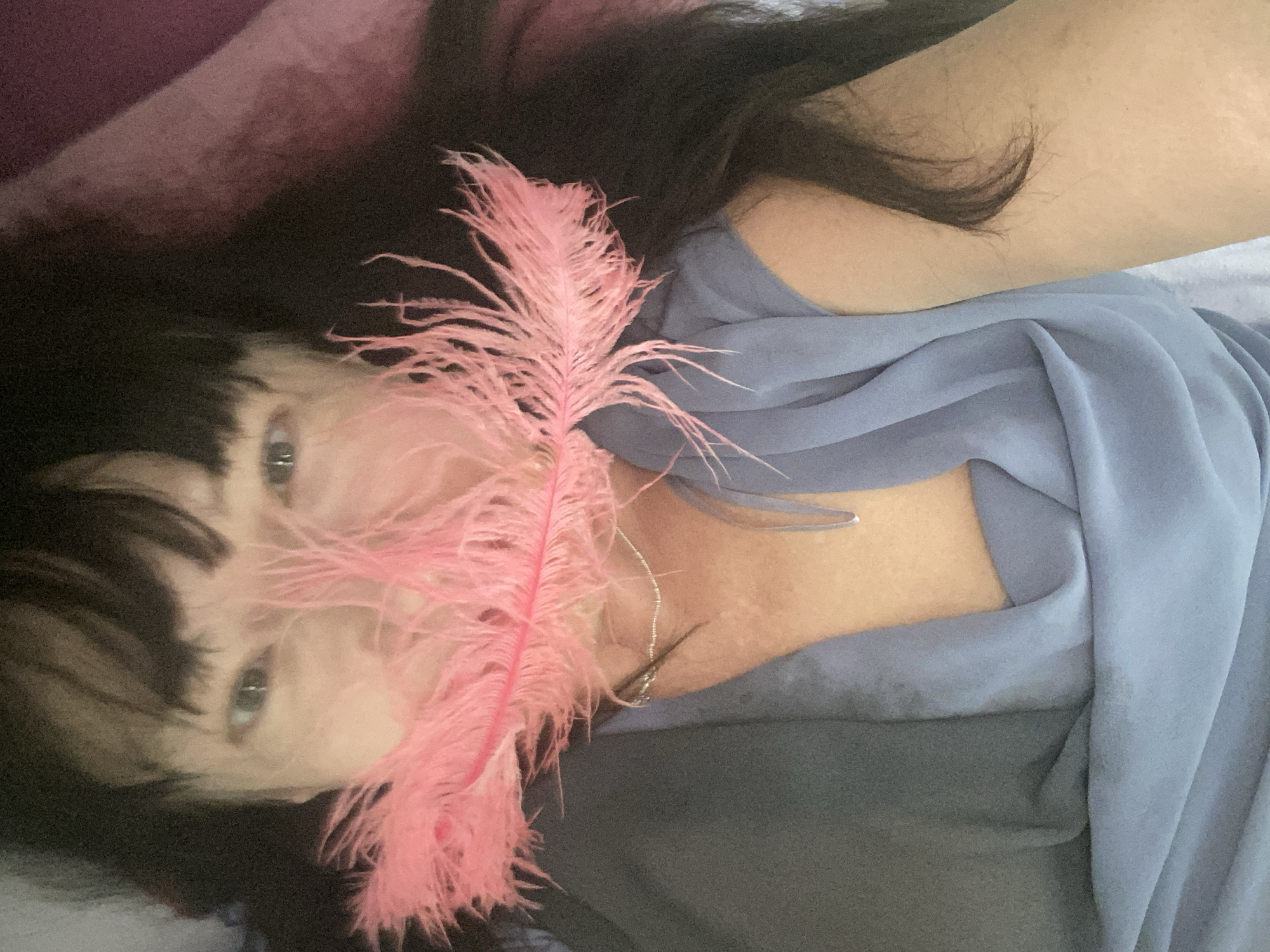 In this close-up, rotated image, a person with dark hair and bangs lies down, their face partially obscured by a fluffy pink feather. The person has light-colored eyes and appears to be looking upwards. They are wearing a light blue, flowing garment and a delicate silver necklace with a small pendant. The background is a mix of dark hair, fabric in shades of purple, blue, and gray, suggesting a comfortable, indoor setting. The lighting appears soft, and there are no visible cues about the time of day or weather. The overall mood is serene and perhaps a bit playful due to the feather. No text is visible in the image.