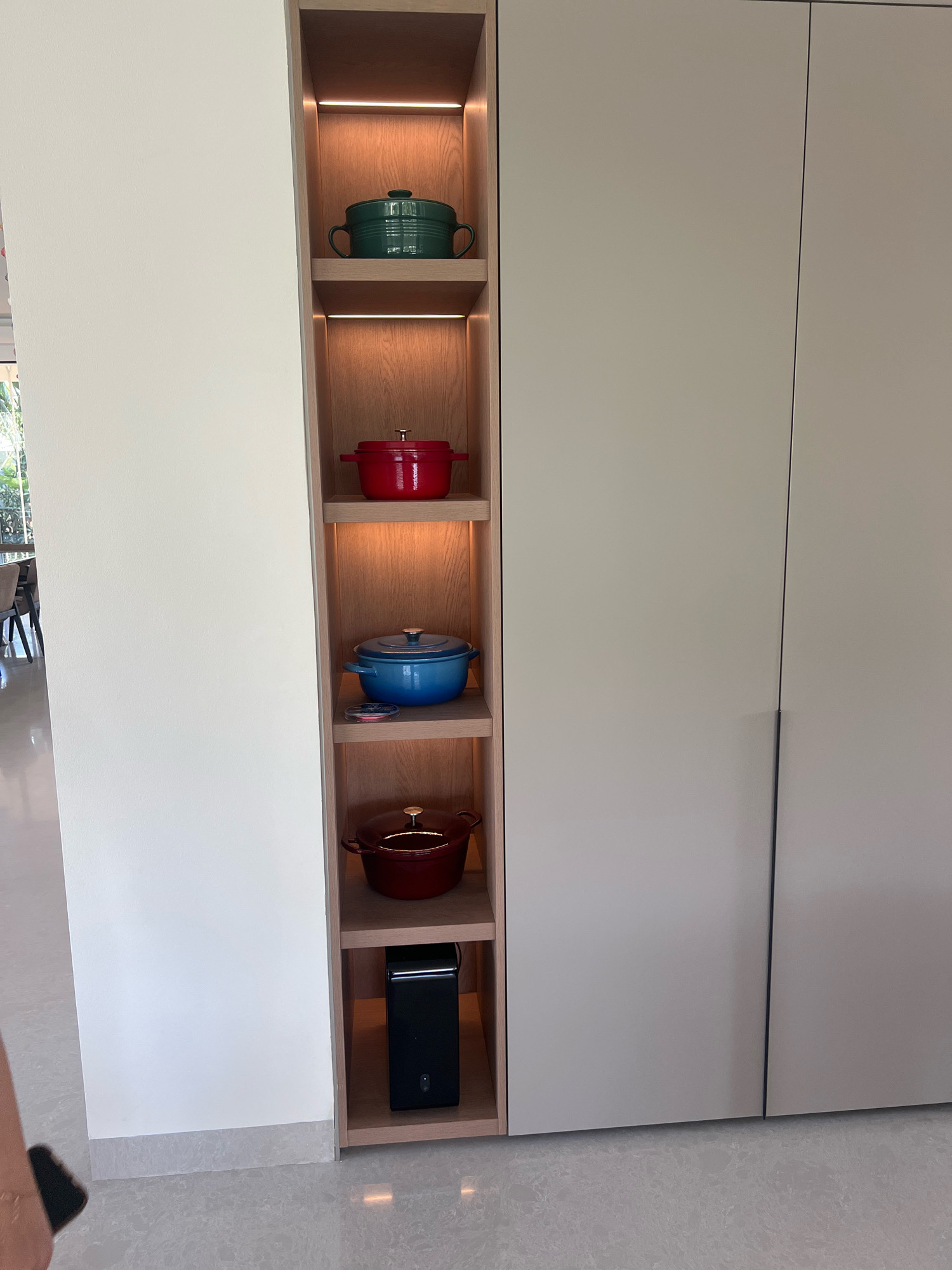 A narrow, tall, built-in shelving unit is positioned against a plain, off-white wall. The shelving unit has wooden shelves that are illuminated by warm LED lights. On the shelves are four enameled cast-iron pots: a green pot on the top shelf, a red pot on the second shelf, a blue pot on the third shelf, and a darker red pot on the fourth shelf. On the bottom shelf of the unit is a small, black electronic device. To the right of the shelving unit are two large, flat, light gray cabinet doors, forming part of a larger cabinetry system. The floor is a light gray, speckled tile. The overall scene appears to be an indoor setting, possibly a kitchen or dining area, in a modern residential or commercial space. The location is identified as Ngaglik, Indonesia, though no specific visual cues in the image confirm this location.