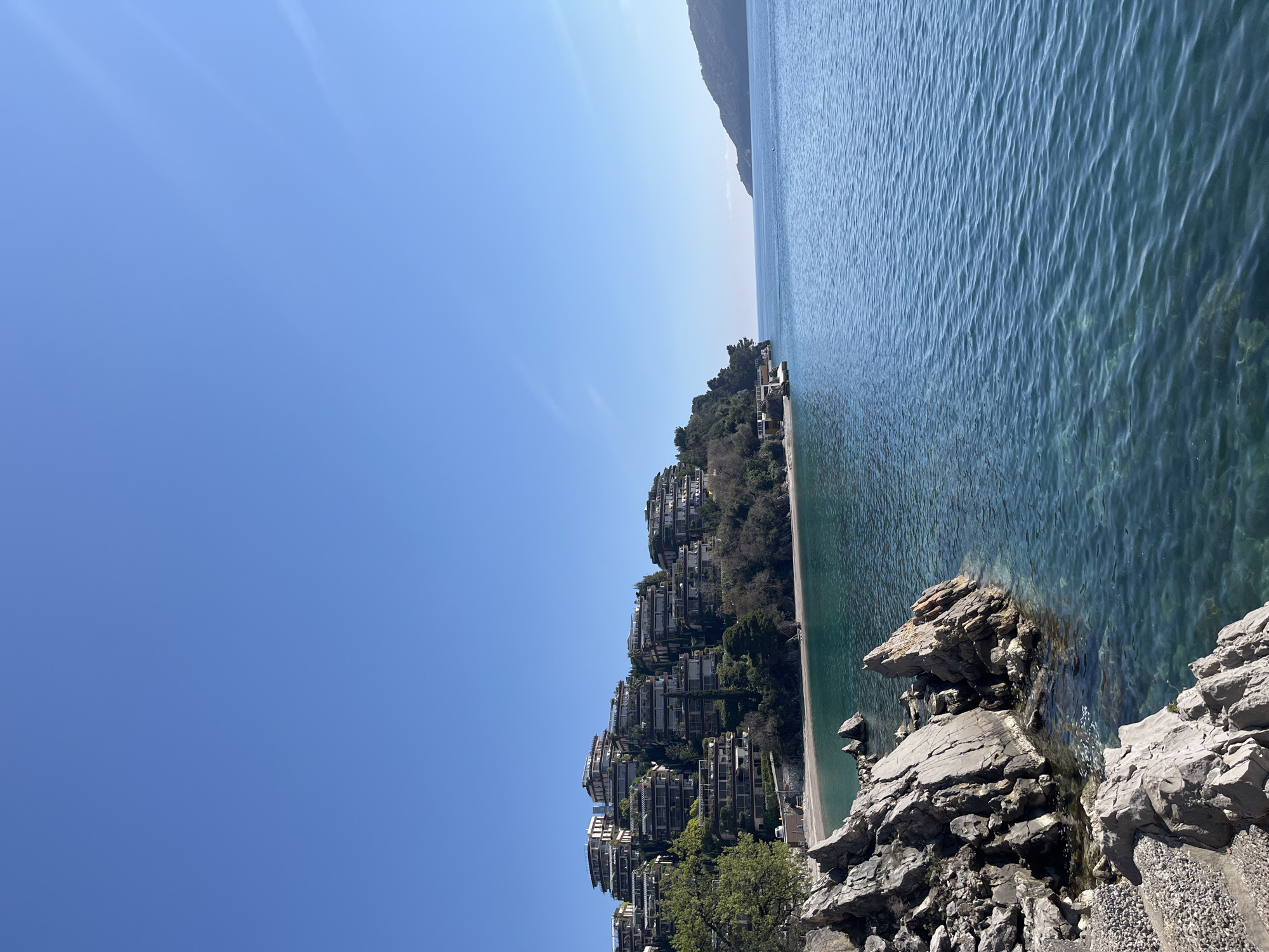 The image shows a coastal scene in Budva, Montenegro. Terraced residential buildings are built on a hill sloping down to the Adriatic Sea. The sea is calm with visible surface ripples. The foreground features rocky outcrops. The sky is clear and blue.