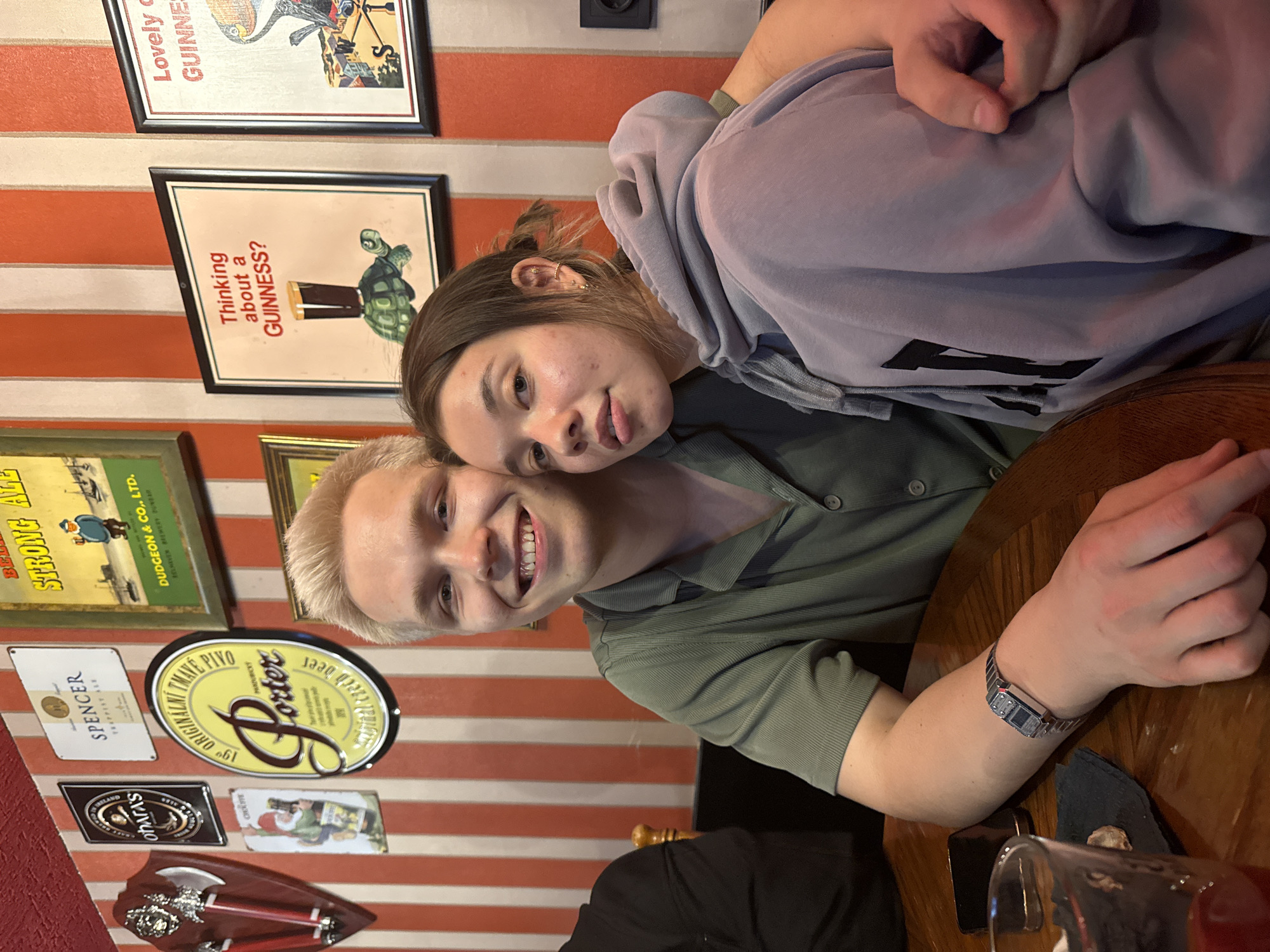Two people, one with blond hair and one with brown hair, are sitting at a wooden table. The person with blond hair is wearing a green shirt and the person with brown hair is wearing a gray hoodie. The table has a glass of liquid on it and a watch. The background has red and white stripes and various signs, including "Lovely GUINNE," "Thinking about a GUINNESS?," "STRONG ALE," and a sign with the word "Porter" on it. Some of the signs are identified as being from Dudgeon & Co., Ltd. Beaver Brewery in Ireland, and "TMAVE Pivo" Pardubicky, chech beer. There is also a sign saying "SPENCER TRAPPIST ALE" and another sign saying "IRELAND OhArA'S CRAFT BEER." The city is Chaikovsky, Russia.