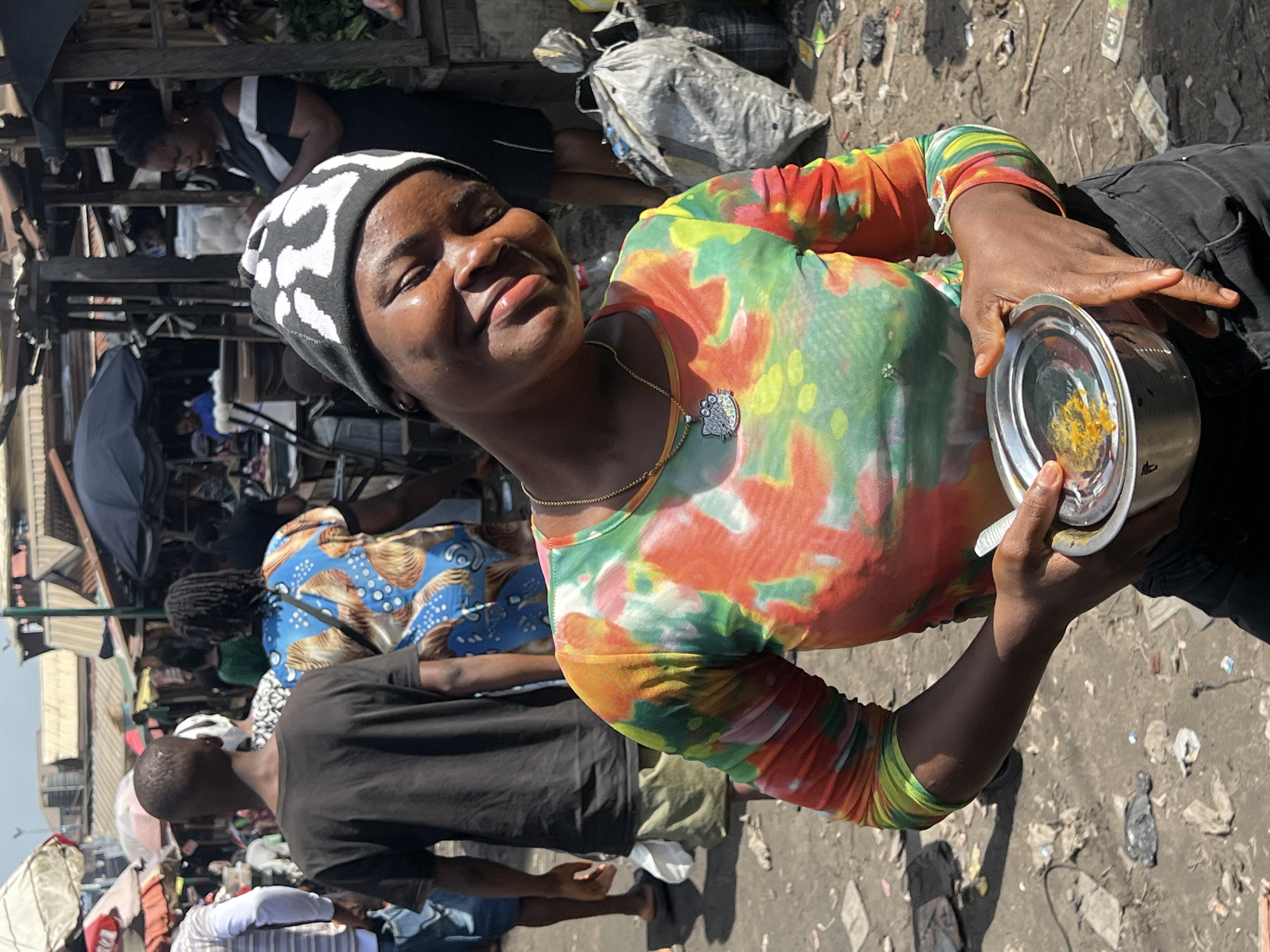 A woman is holding a metal bowl of food in a crowded outdoor market. The woman, who is the central figure, is wearing a tie-dye shirt and a black and white beanie. She is also wearing a gold chain with a pendant. The bowl contains a yellow food item.
The background indicates a bustling market scene, likely in Nkpor, Nigeria. There are several people around, including other market vendors and customers. The ground appears to be littered with debris. Various structures, umbrellas, and stalls are visible in the periphery, typical of a market environment.