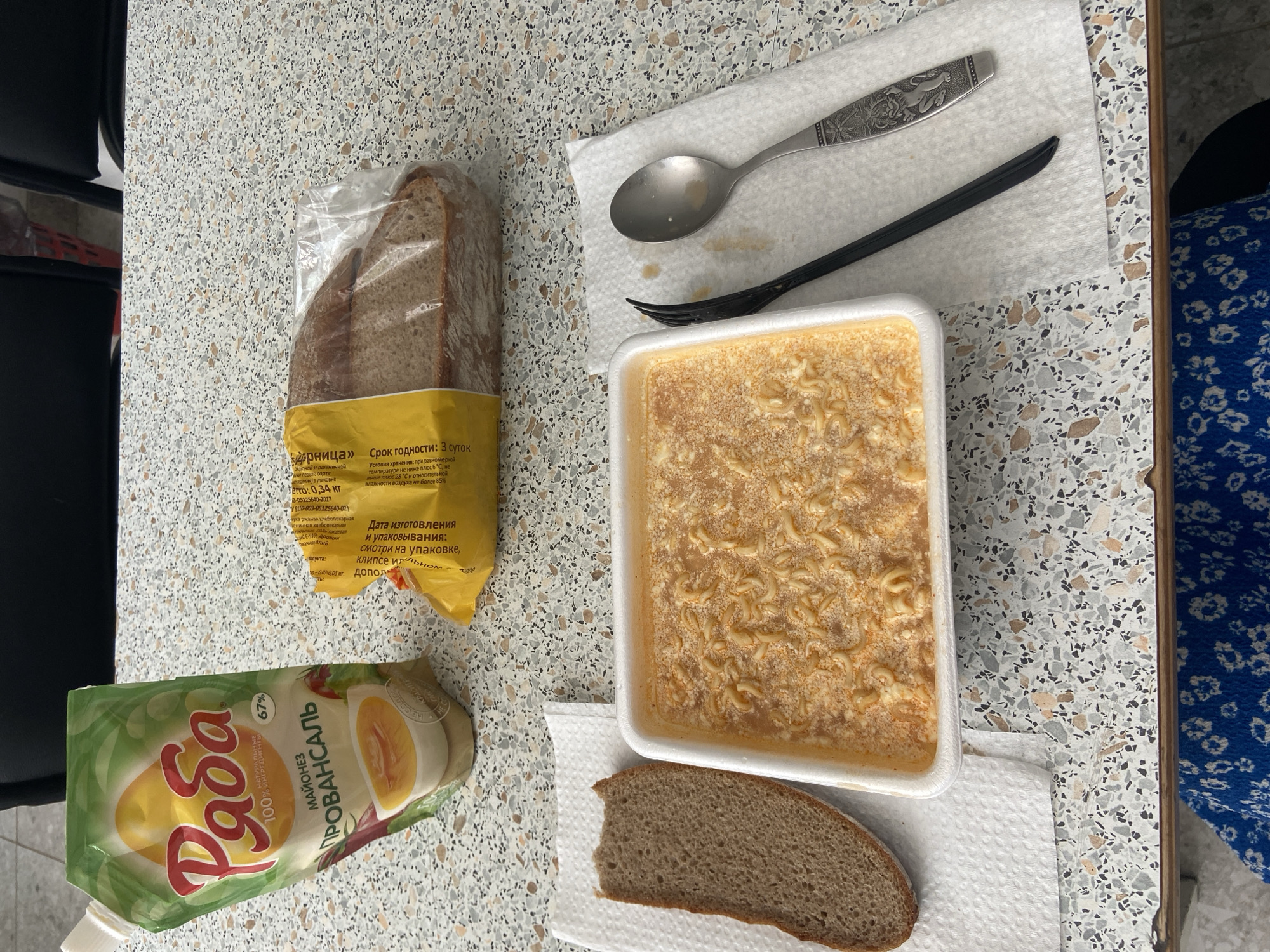 On a countertop with a terrazzo pattern, likely in a home setting, is a meal laid out, consisting of: a sealed bag of sliced brown bread; a packet of "Ryaba" brand mayonnaise, labeled "Provansal"; a white plastic container holding noodles in a broth; a single slice of brown bread on a napkin; and a metal spoon and black plastic fork on a separate napkin. The packaging on the bread has Russian text indicating the bread is called "Udarnitsa" and has a 3-day shelf life. Based on the Cyrillic text on the food packaging, the location is likely in Russia. Given the brand of mayonnaise, it could specifically be located in the city of Chaikovsky, Russia, where the manufacturer is based. A black chair is visible on the left and the edge of a blue flowered tablecloth is visible on the right.