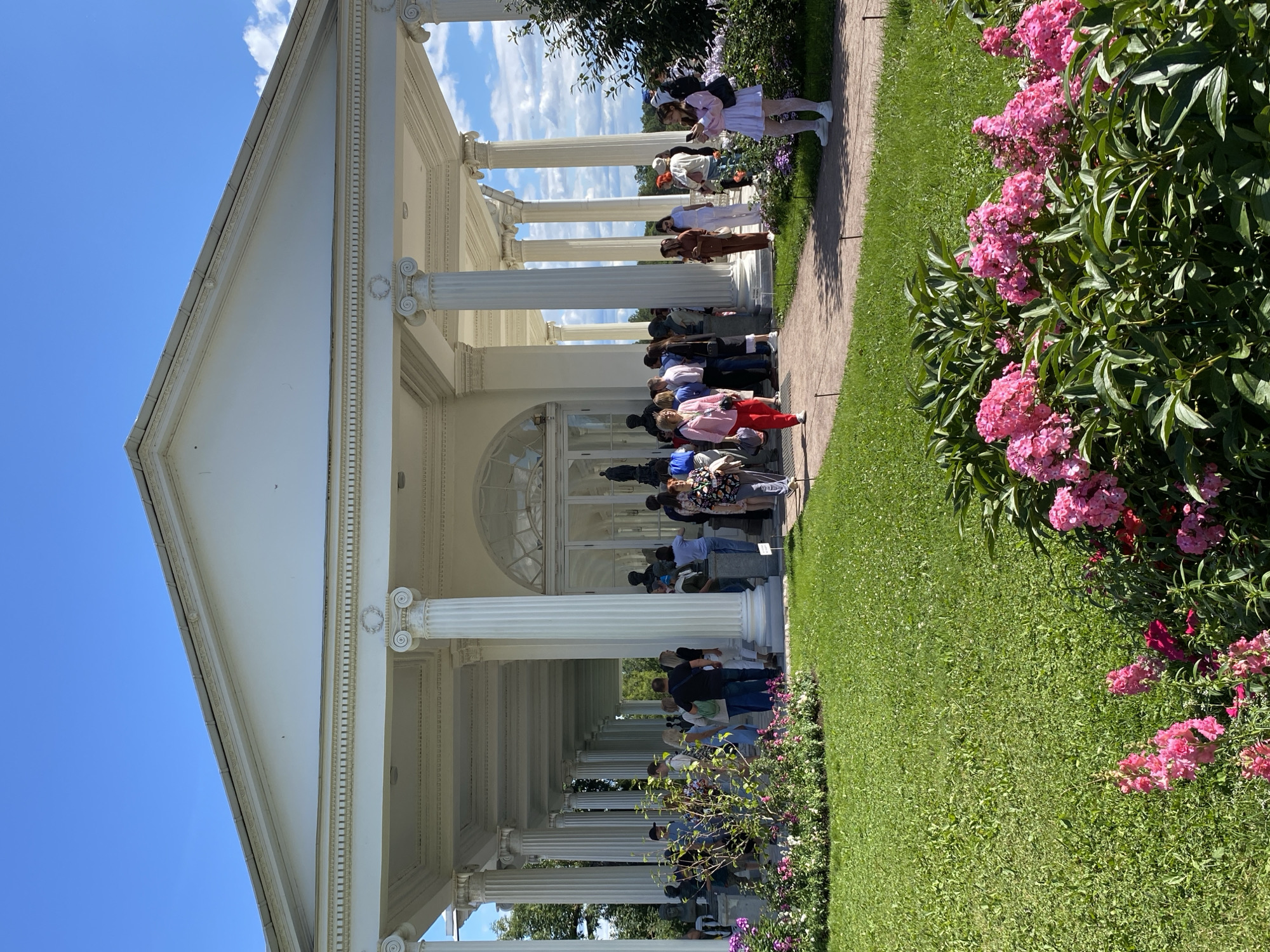 A wide-angle, eye-level shot captures a sunlit, manicured garden with a large, white neoclassical building in the background. The building features a long, colonnaded portico with prominent ionic columns and a triangular pediment. A crowd of people, dressed in casual summer attire, are gathered on the paved walkway in front of the building, some appearing to be observing sculptures or exhibits inside. The foreground is dominated by lush green grass and vibrant pink flowers. The sky is a clear blue with scattered white clouds, indicating a pleasant day. The architectural style and the presence of statues suggest this location is likely a museum or a historical estate. Based on the architecture and the surrounding landscape, the location is identified as the Agate Pavilion in Catherine Park, Pushkin, Russia.