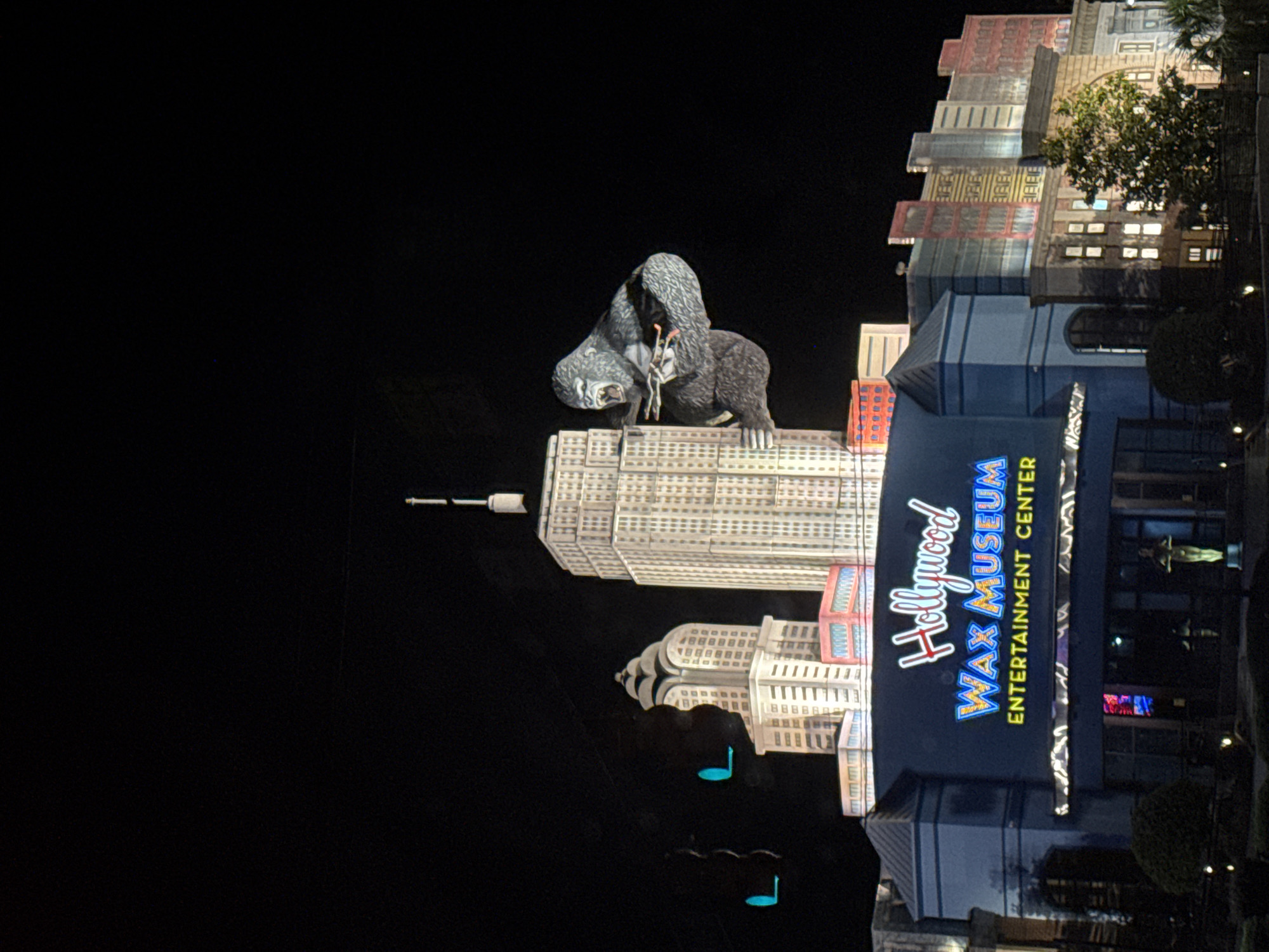 The image shows the Hollywood Wax Museum Entertainment Center in Myrtle Beach, South Carolina, United States. The museum has a large sign with the words "Hollywood Wax Museum Entertainment Center." Atop of the museum, a large mechanical gorilla is perched atop a replica of the Empire State Building. The photo was taken at night.