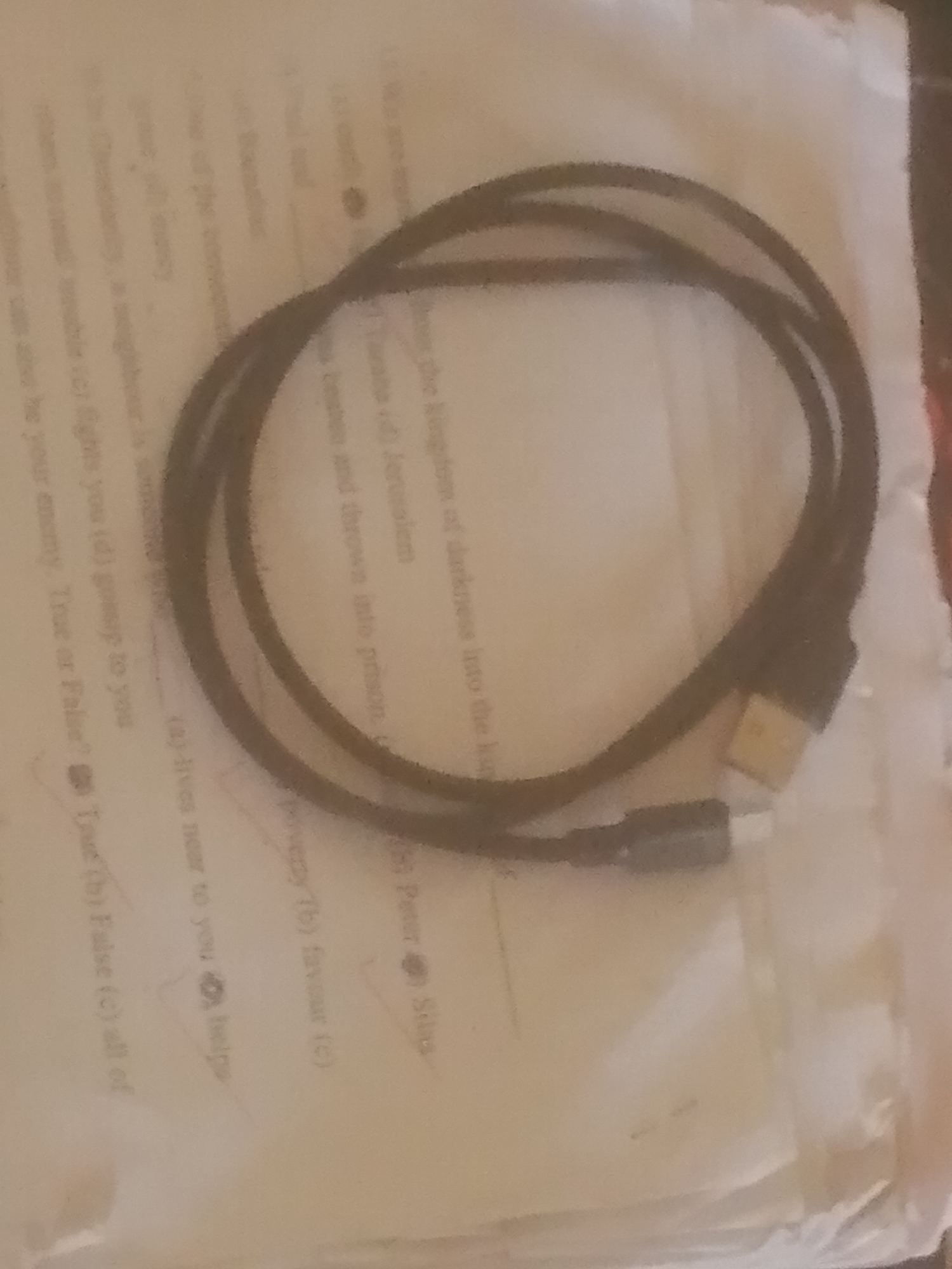 The image shows a black cable with USB connectors, lying on top of a document. The document appears to be a printed paper with text and multiple-choice questions. The text is partially obscured by the cable and the lighting conditions, but some phrases like "kingdom of darkness," "Jerusalem," and "True or False?" are visible. The background is a blurry white surface, possibly a table or a book. There is no indication of a specific location in Jauro Yinu, Nigeria, within the image.