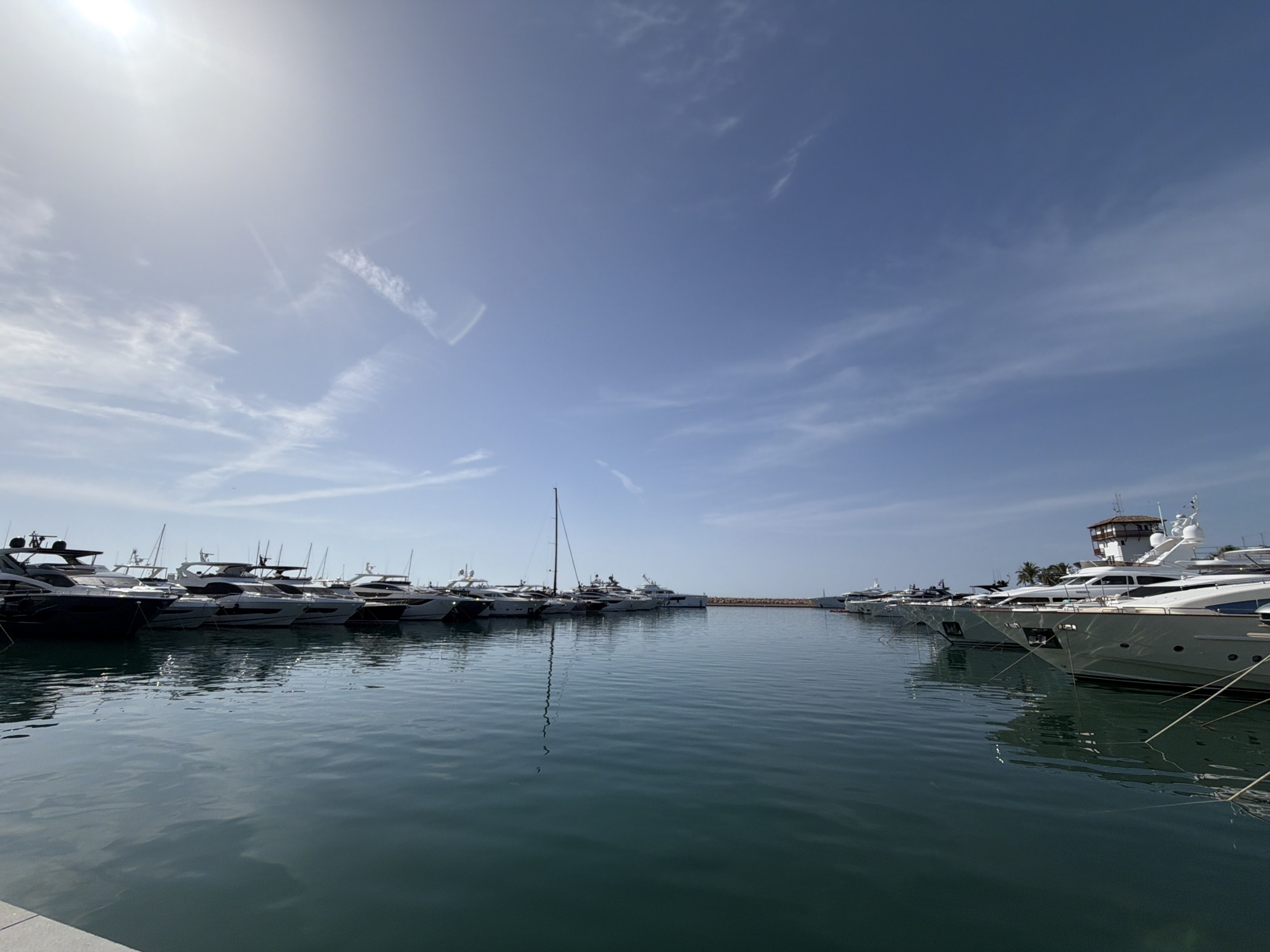 The image depicts a marina filled with numerous luxury yachts docked in calm, turquoise waters. The sun is bright, creating a glare in the upper left corner of the sky, which is a clear blue with wispy white clouds. The yachts are lined up along the pier, with their reflections visible in the water. On the right side of the image, a particularly large, white yacht with multiple levels and a captain's tower is prominently featured, its hull shining in the sunlight. Beyond the yachts, a breakwater or pier structure can be seen in the distance. Palm trees are visible further back, suggesting a warm climate. The scene is peaceful and serene, conveying a sense of opulence and leisure associated with yachting. The location is Portals Nous, Spain.