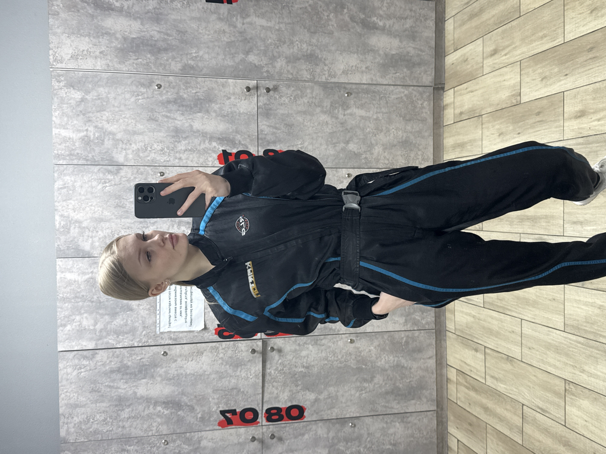 A young Caucasian woman with blonde hair is standing inside a locker room in Perm, Russia. She is wearing a black racing suit with blue trim and a buckle around the waist. She is holding a black smartphone in her left hand and is taking a selfie. The lockers are gray with black and red numbers affixed to them. The floor is tiled, with the lockers on the left side of the image and the tiled floor on the right. There is a printed paper attached to one of the lockers.