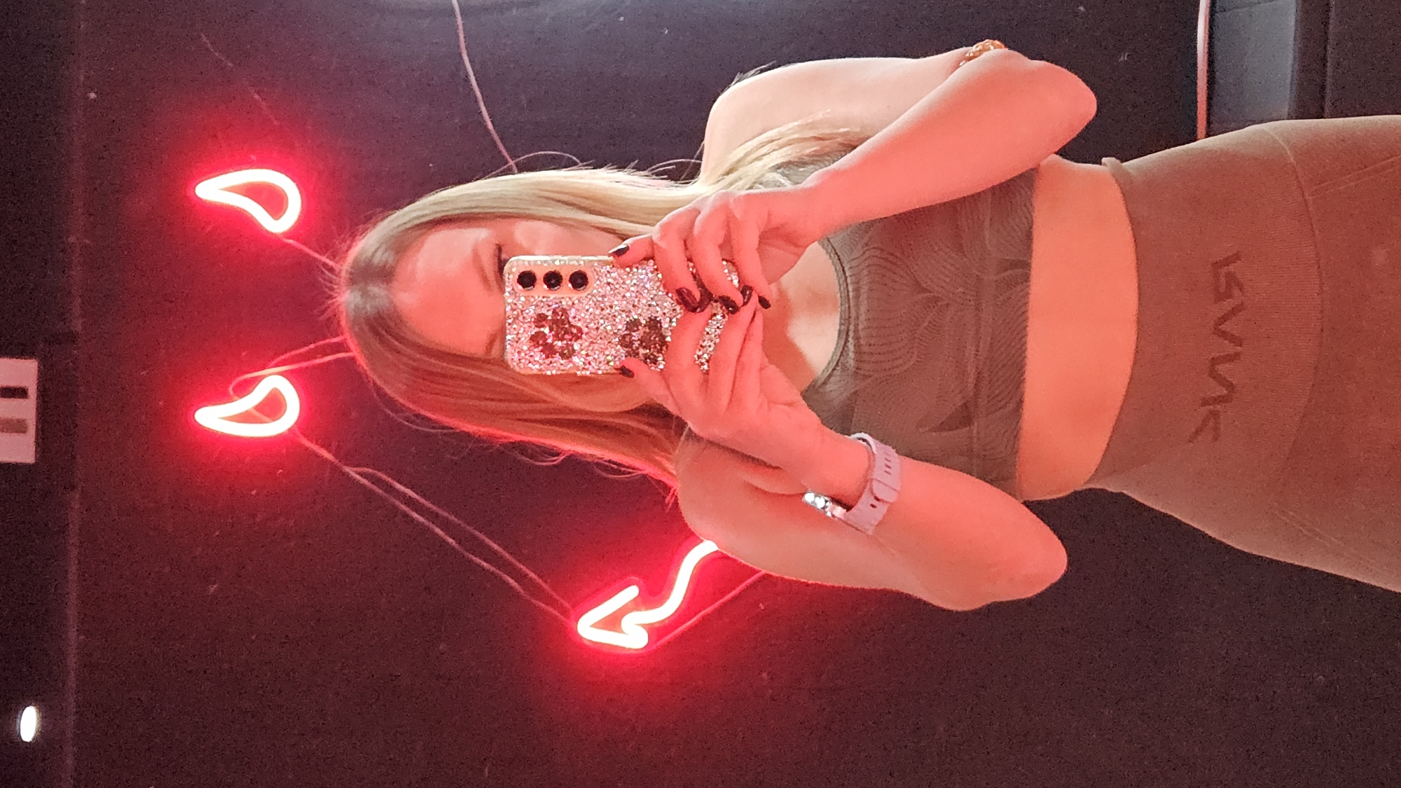 A person is taking a selfie in front of a red neon light fixture resembling devil horns and an arrow. The person is wearing a matching top and leggings outfit. The person is holding a phone with a rhinestone-covered case. The location is not identifiable as the city of Sevastopol, Ukraine.
