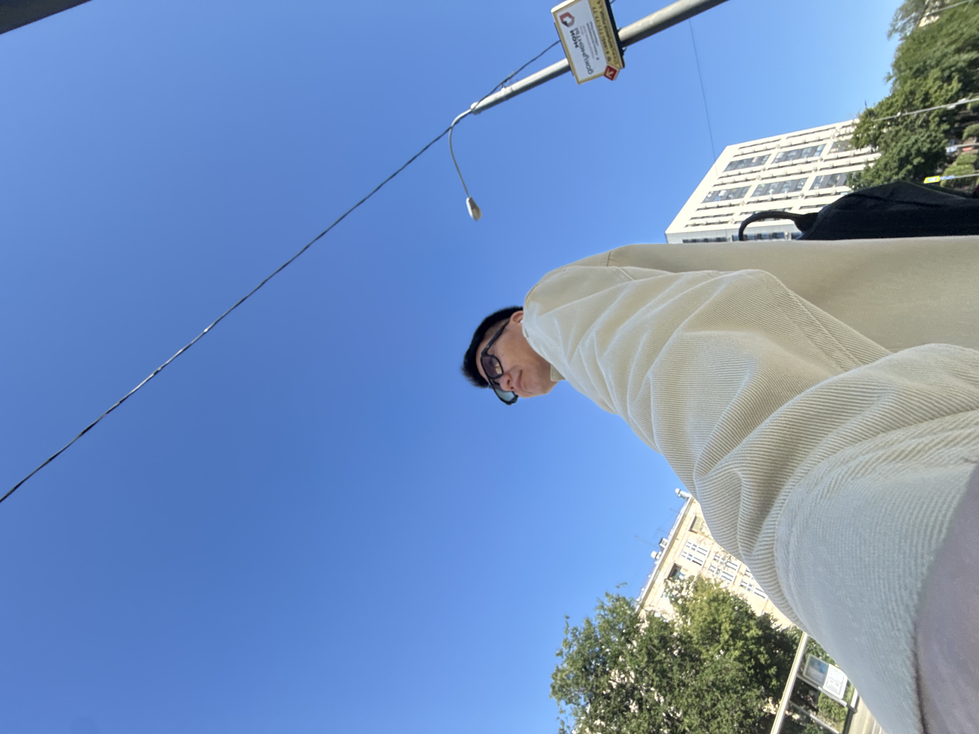An eye-level, low-angle outdoor medium shot captures a person wearing a light-colored corduroy jacket and dark sunglasses against a bright blue sky. The person is looking upwards and to the right. A black umbrella is visible behind their shoulder. In the background, a tall, modern white building with numerous windows stands to the right, partially obscured by trees. To the left, another building with a lighter facade and many windows is visible, along with dense green trees and a bus stop structure. A lamppost with a sign is positioned to the right of the person, and a wire runs diagonally across the upper portion of the frame. The image appears to be taken on a sunny day in Moscow, Russia, given the context of the provided hint.