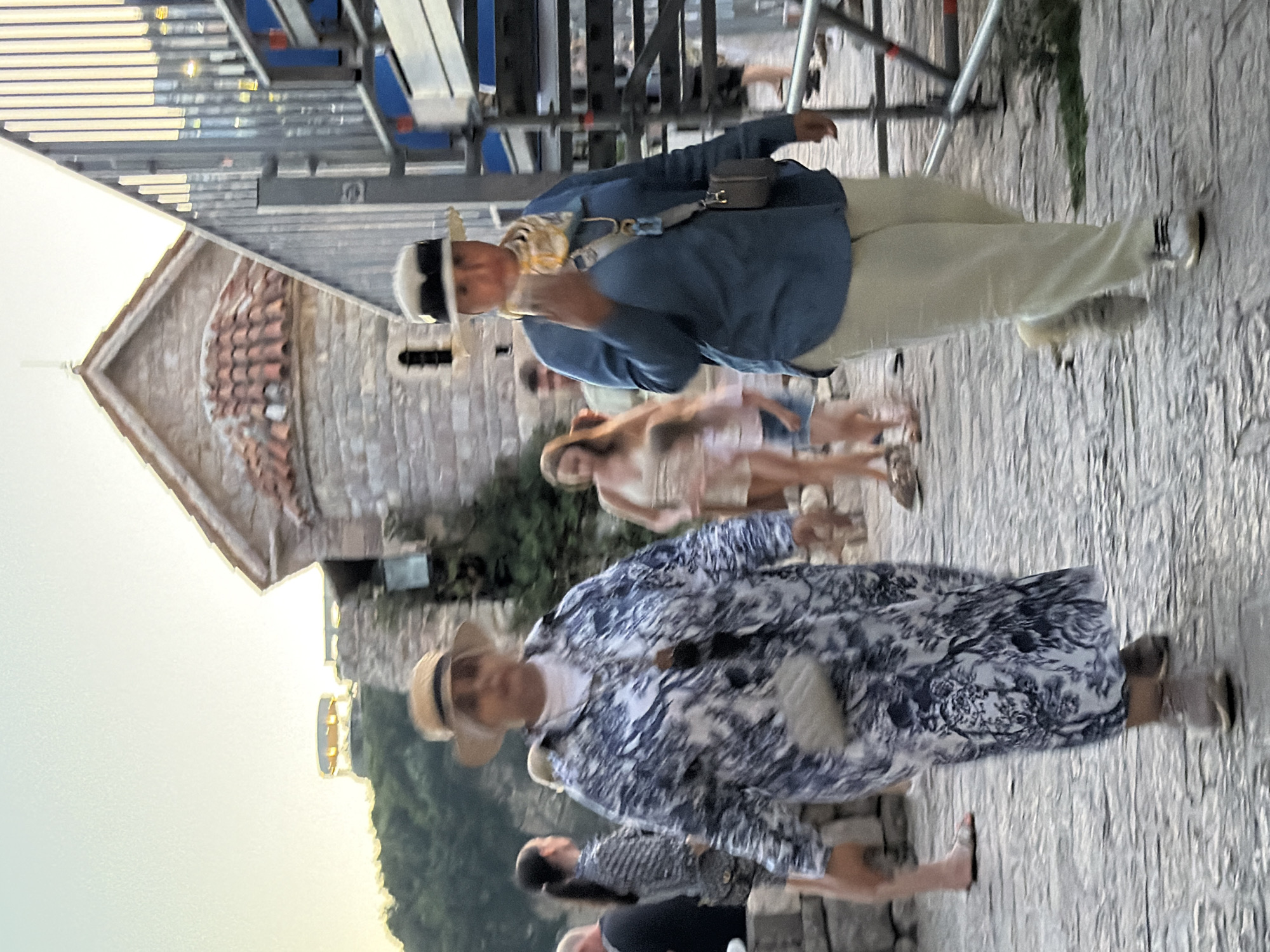 In Budva, Montenegro, two women walk on a cobblestone path near an ancient stone building with a red-tiled roof. The woman on the left wears a blue and white patterned long coat, a white shirt, a straw hat, and light-colored shoes. She carries a small quilted crossbody bag. The woman on the right wears a blue long-sleeved shirt, light-colored pants, a straw hat with a dark band, a patterned scarf, and white sneakers. She holds a phone to her ear and has a dark crossbody bag. In the background, other people are visible, some walking and others standing. To the right, a modern metal structure with blue accents is under construction or being set up, contrasting with the historical architecture. The sky is bright and hazy, suggesting late afternoon or early evening.