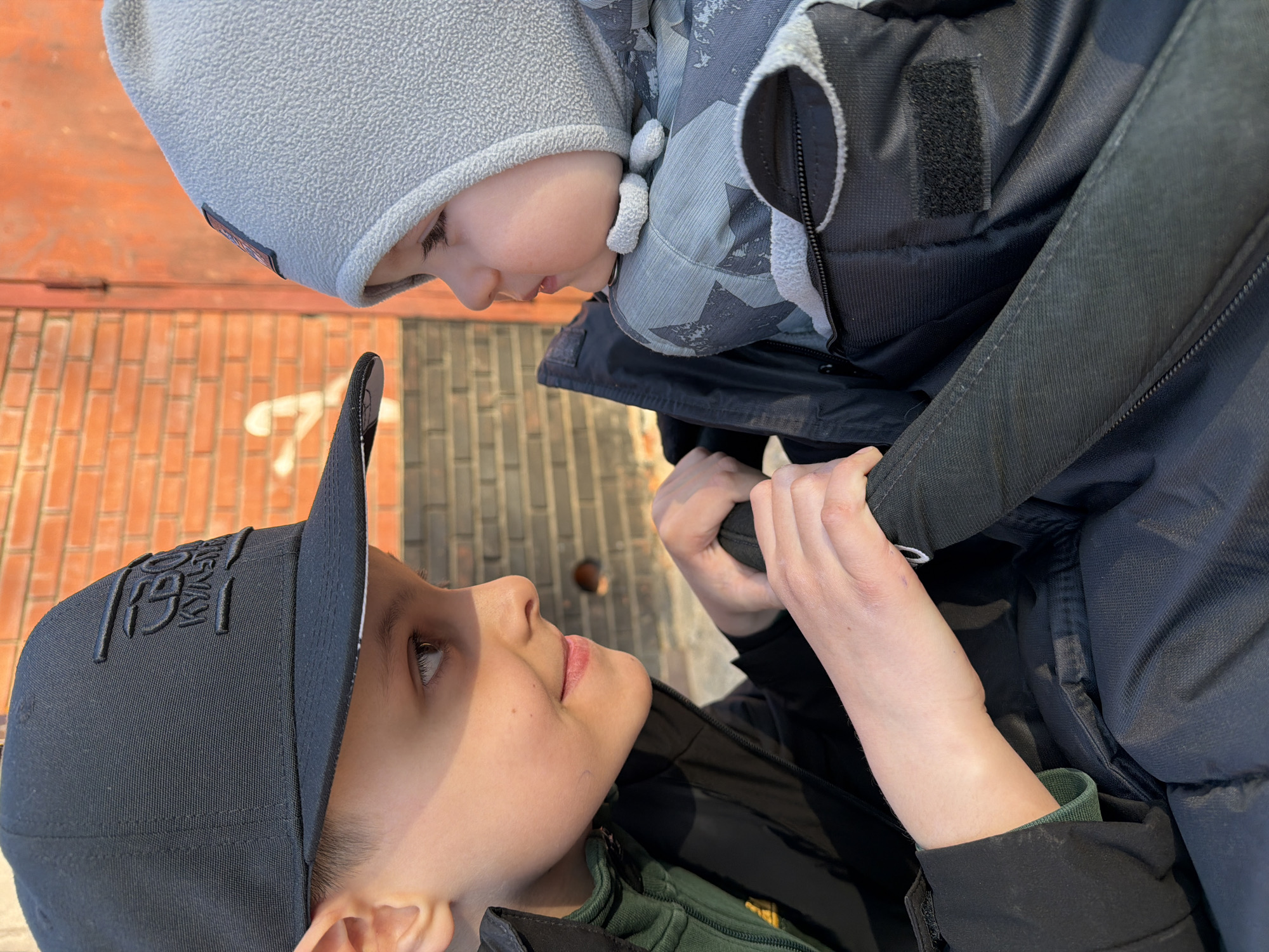 This image captures an intimate moment between two children, an older boy and a younger infant or toddler. The photograph is oriented sideways, depicting the older boy looking up at the younger child, who is positioned above him and looking down.
The older boy is wearing a black baseball cap with embossed text, which appears to be Cyrillic, possibly reading "ВОТ КУЛЬТ" (VOT KULT). He is also wearing a dark green long-sleeved shirt and a black jacket. His face, seen in profile, shows a gentle and focused expression as he gazes at the younger child. His hands are visible, gripping the younger child's outerwear, suggesting he is holding or supporting them.
The younger child is dressed in a light grey fleece hat and a grey and black patterned winter jacket or carrier. Their face, seen from the side, exhibits soft features and a calm expression as they look down towards the older boy.
The background is a textured red-brown brick wall, indicating an outdoor setting in Votkinsk, Russia. The scene is brightly lit by strong sunlight, casting visible shadows and highlighting the children's features, suggesting a clear and sunny day. The children's clothing implies cooler weather. The interaction between them conveys a tender and affectionate connection.